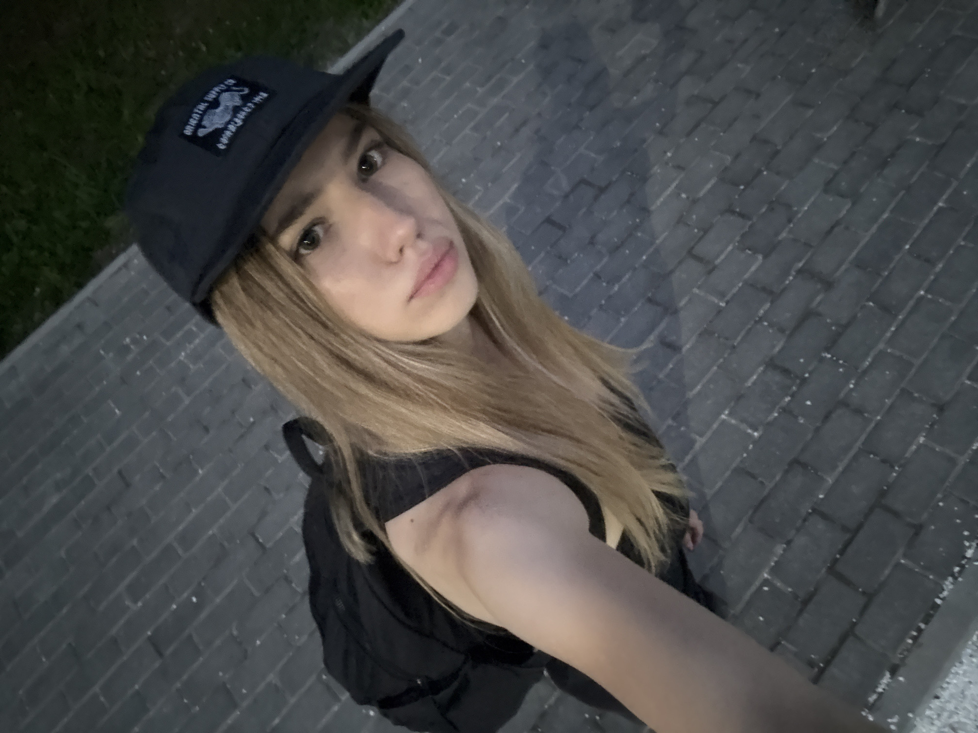 A young woman with long blonde hair is shown in a selfie taken outdoors at night. She is wearing a black baseball cap with a patch that has a leopard design, and a black sleeveless top. Her backpack is visible. The background consists of a paved area with gray interlocking bricks, and some green grass on the left. The scene appears to be in an urban setting, possibly a park or a street in Chaikovsky, Russia.