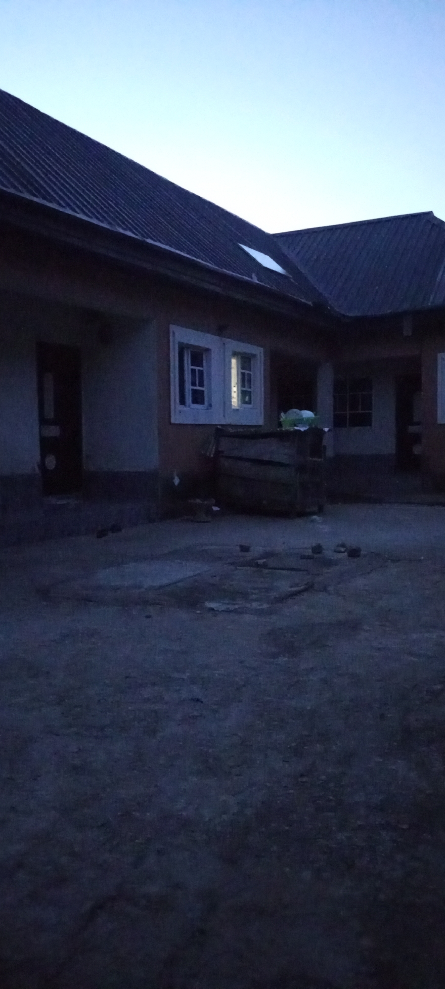 The image depicts a courtyard area in front of a building with a corrugated metal roof. The sky above is a pale blue, indicating either dawn or dusk. The building has several windows, some of which are lit from within. In the foreground, the ground is paved with rough, uneven material. A wooden structure, possibly a crate or a makeshift bench, is visible near the building. The overall scene suggests a residential or possibly a small commercial property in an urban setting. The lighting and atmosphere imply a quiet time of day. The location is identified as the city of Jalingo, Nigeria.