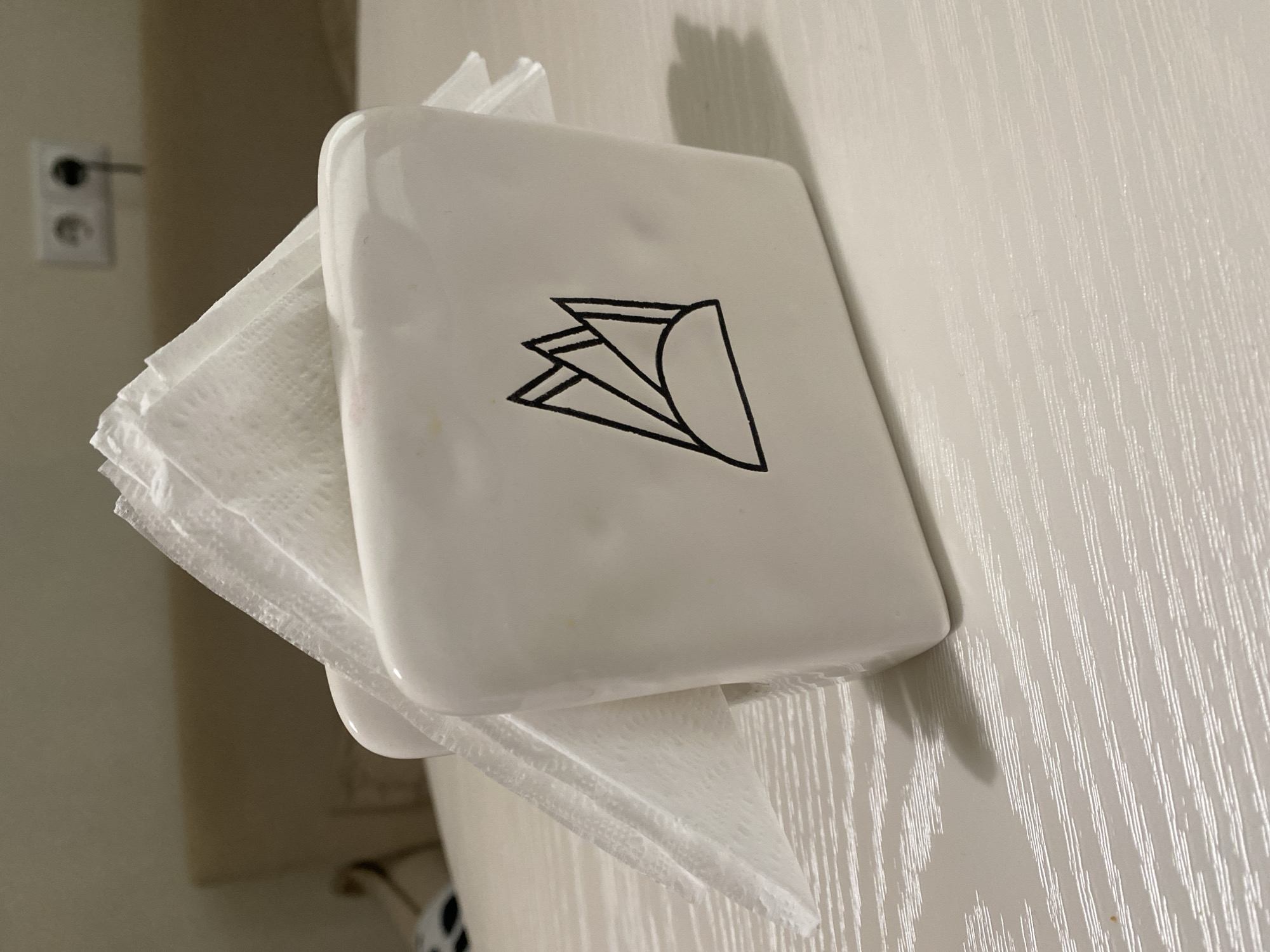 The image depicts a white, glossy ceramic or plastic napkin holder filled with multiple white paper napkins, positioned centrally on a light-toned, horizontally textured surface. The scene takes place in an indoor setting in the city of Unknown, Unknown.
The napkin holder displays a minimalist black line-art design on its visible front face, illustrating a stylized representation of folded napkins with geometric peaks above a curved base. Several white paper napkins, exhibiting a slight texture, are folded and protrude from the top opening of the holder.
The surface beneath the holder, likely a table, is light-colored and features a distinct horizontal grain or striation pattern. A clear shadow of the napkin holder is cast to its right, indicating a light source from the left.
In the out-of-focus background, a light-colored wall is visible. A blurry, light brown upholstered object, possibly a headboard or sofa back, extends horizontally across the mid-ground. In the upper right portion of the background, a white double electrical outlet is mounted on the wall; its right socket appears to have a dark plug or object inserted. A small, dark object with a light pattern, possibly part of a cushion or textile, is partially visible in the lower-left background. No people, actions, or interactions are present in the image.