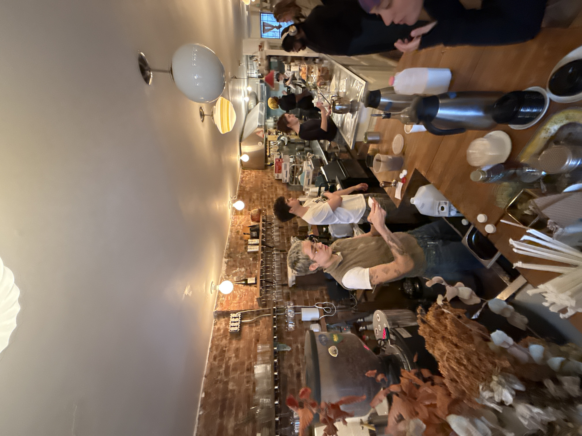 The image shows an interior view of a cafe or restaurant in New York, United States. Patrons stand along a counter, seemingly interacting with the staff. The scene is illuminated by overhead lighting fixtures, and the architecture includes brick walls on one side, suggesting an urban setting. A variety of equipment and utensils are present on the counter, indicative of a working service area. The perspective is slightly tilted.
