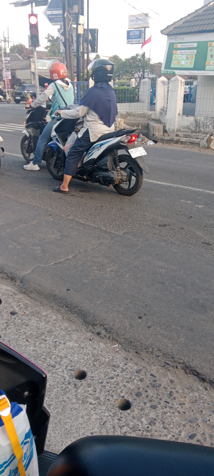 In the city of Maguwoharjo, Indonesia, two individuals are riding on a motorcycle at a traffic intersection. The person in the front wears an orange helmet and a light blue jacket over a white shirt. The person in the back has a dark blue hijab and a white and black striped long-sleeved shirt. They are both looking forward. The motorcycle is white and blue, with a license plate that reads "AB 5375 TD". To the left of the motorcycle, a traffic light shows a red signal. In the background, there are buildings, including a bank with an ATM, and trees. The road surface is asphalt.