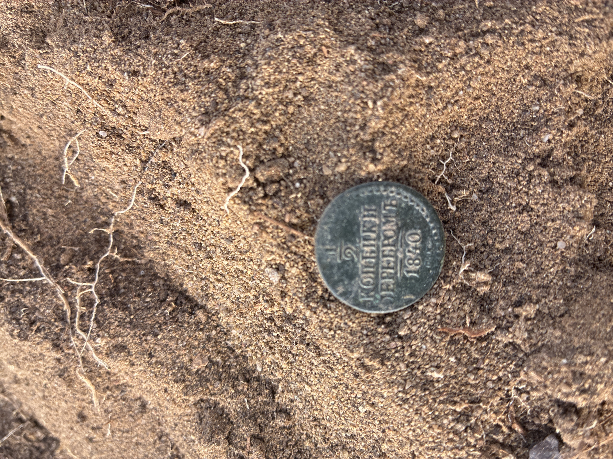 A close-up, overhead shot shows a single, dark-colored coin partially embedded in sandy soil. The coin, which appears to be made of copper or bronze, has visible inscriptions on its surface. The most legible text reads "1840" at the bottom, above which is a fractional symbol and what appears to be Cyrillic script. The coin is situated in a patch of disturbed earth, with several thin, dry roots or plant fibers scattered across the surface. The background is filled with the same granular soil, with subtle variations in color and texture. The lighting suggests it is daytime, with a warm, natural light casting soft shadows. The location is identified as Ust'-Ordynskii, Russia, though no specific landmarks or features of the city are visible in the frame.
