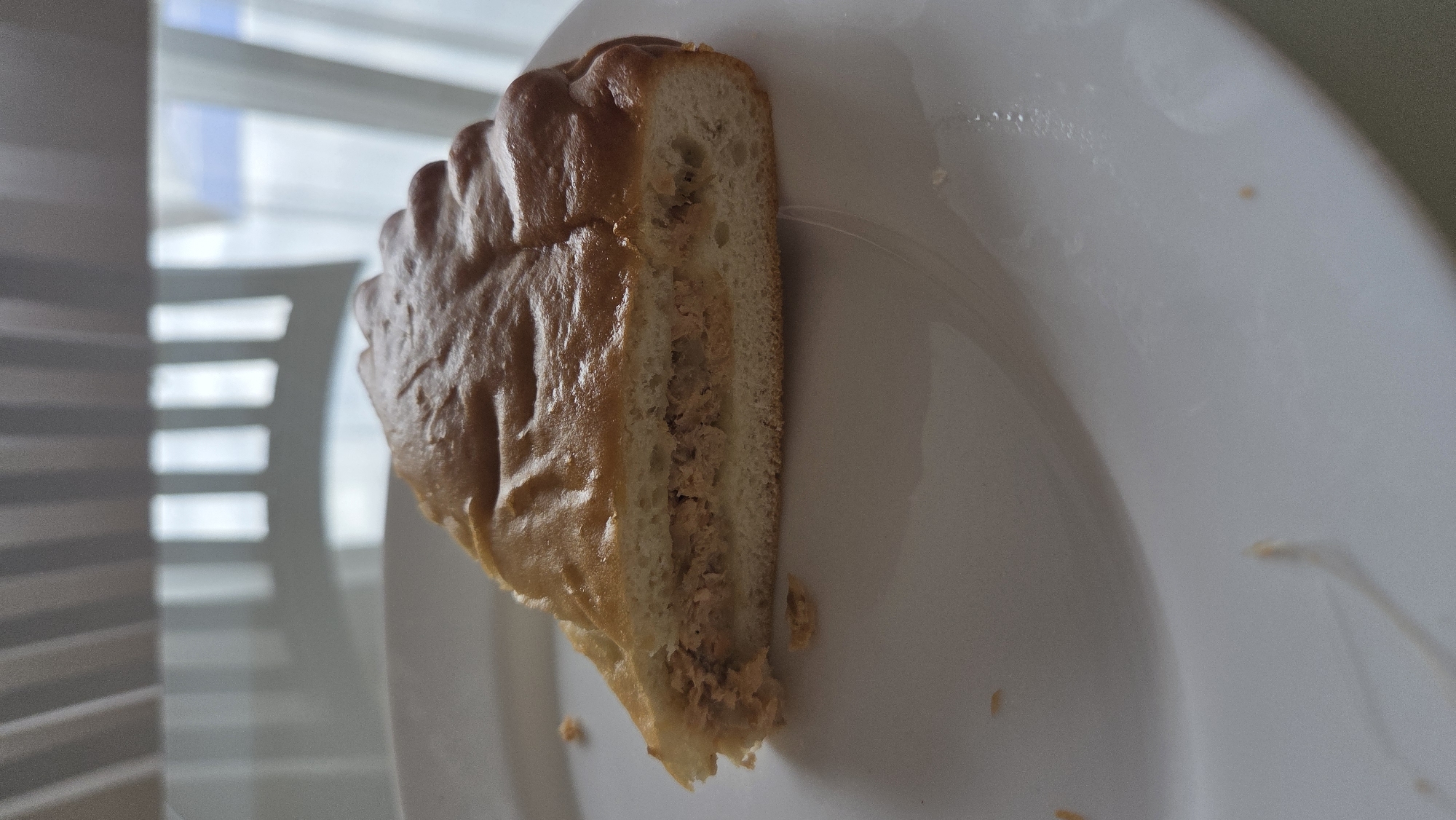 A close-up, slightly angled shot shows a slice of a golden-brown baked good resting on a plain white plate. The pastry, possibly a type of pirog common in Russia, features a rich, slightly glossy crust with subtle decorative folds or seams on its upper surface. The exposed cross-section reveals a light brown, finely textured filling, suggesting it could be savory, perhaps meat or fish, encased within the soft dough.
The white plate appears largely clean, though a few small crumbs or pieces of the filling are visible, indicating the pastry has been cut or partially consumed. In the background, out of focus, is a window with horizontal blinds, allowing natural daylight to filter in. The strong light creates distinct horizontal shadow patterns from the blinds on what appears to be an interior wall or another surface, suggesting an indoor setting during the daytime. No people are visible, and no text is discernible. The scene implies a simple, everyday moment, likely in a home or casual eating establishment, in a location such as Chaikovsky, Russia.