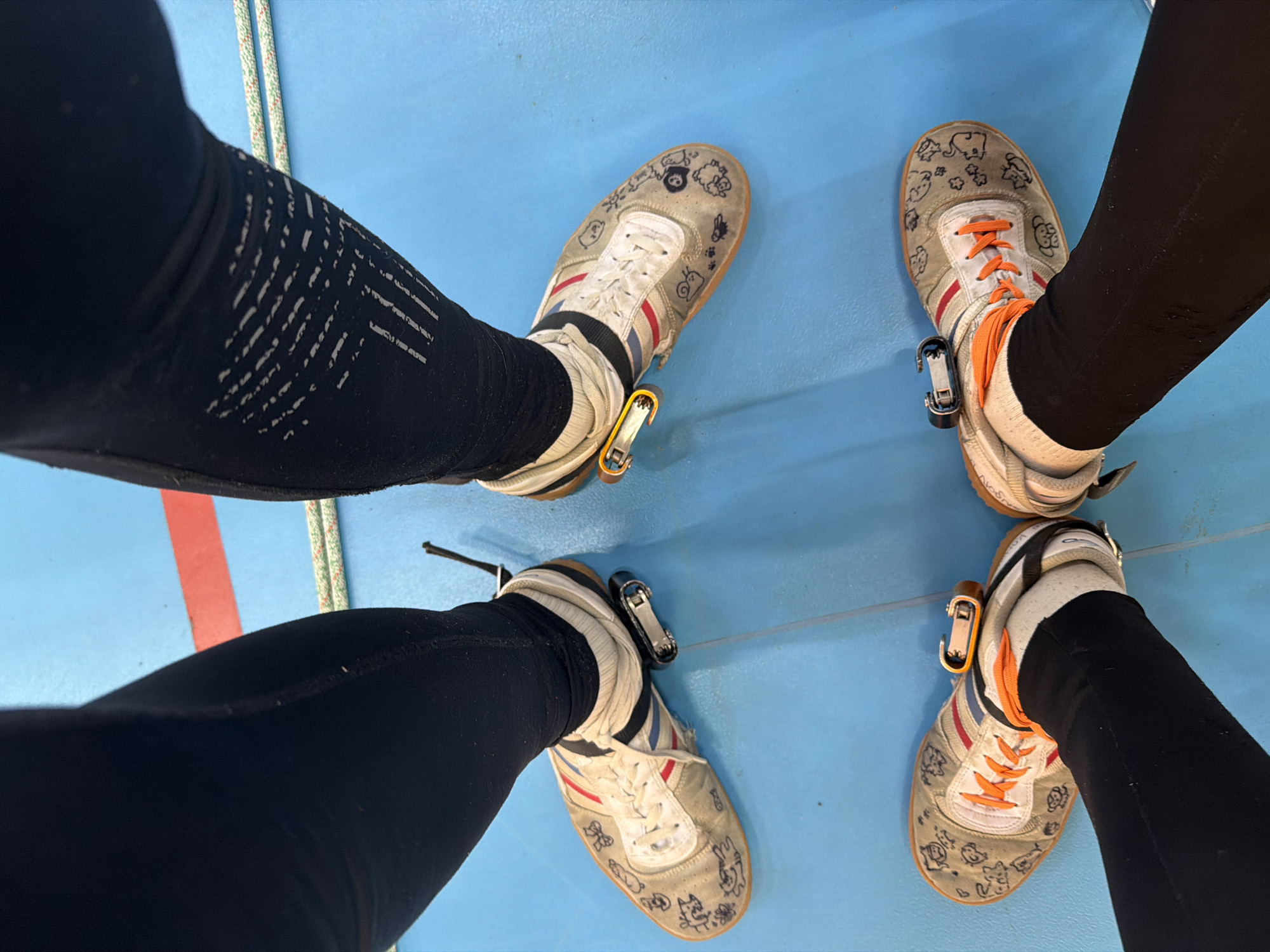 The image shows the lower legs and feet of two individuals standing on a light blue surface. Both individuals are wearing black leggings and sneakers. The sneakers are similar in style and appear worn, decorated with hand-drawn cartoon characters. Metal devices are attached to the laces of each shoe. A red stripe is visible on the blue surface, and a section of rope is visible in the upper left corner. The location is presumably an indoor recreational or athletic space. It is unknown if this is the city of Berezniki, Russia.