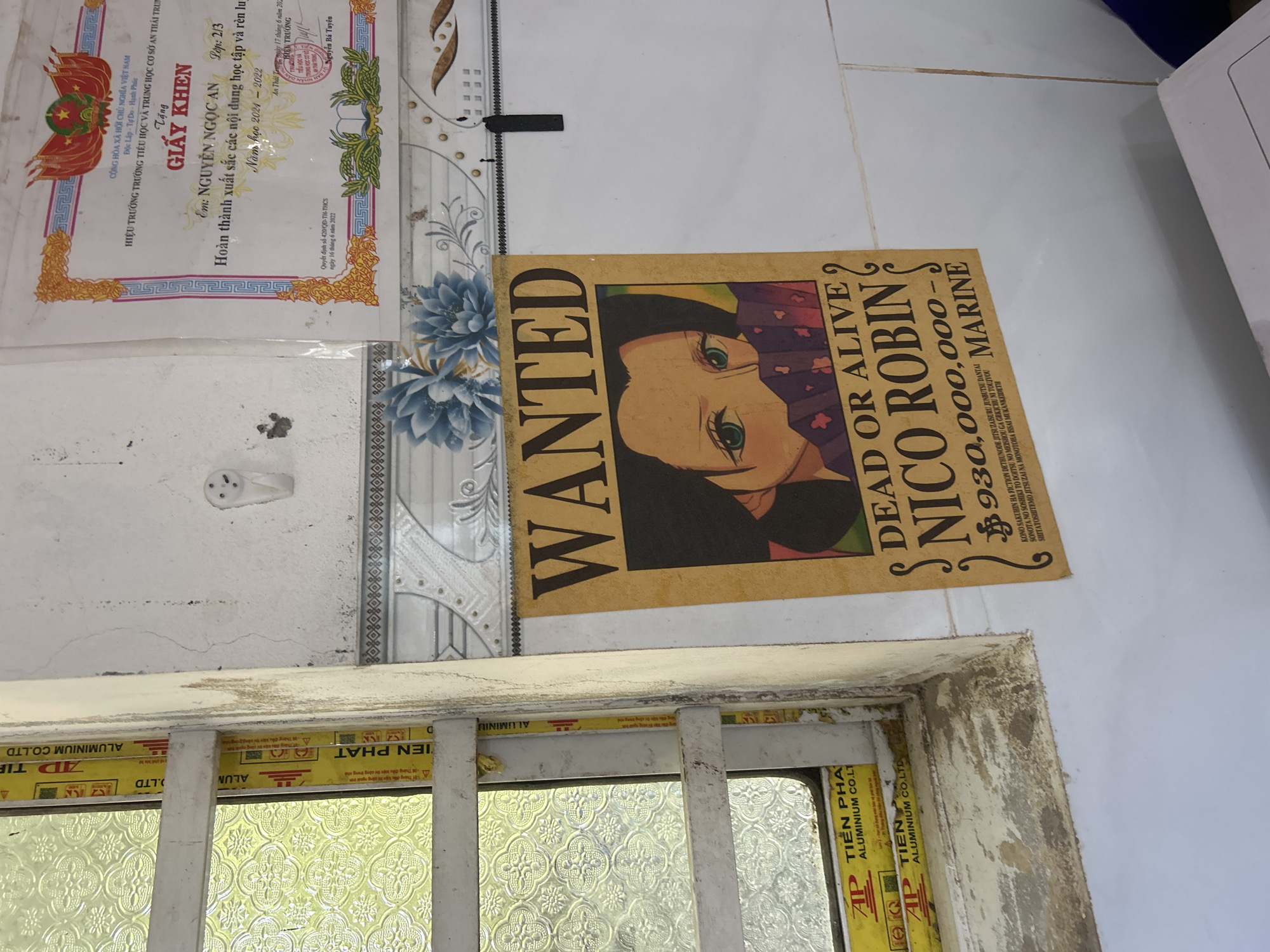 The image shows a poster of Nico Robin from One Piece that is taped to a wall. The poster is yellow and features the words "WANTED" and "DEAD OR ALIVE" along with a picture of Nico Robin and a bounty of 930,000,000-MARINE. The poster is hanging on a tiled wall, and there is a window to the left of the poster. A certificate is also visible on the wall above the poster. The room appears to be dimly lit and may be an office or classroom. The location is identified as An Hữu, Vietnam.