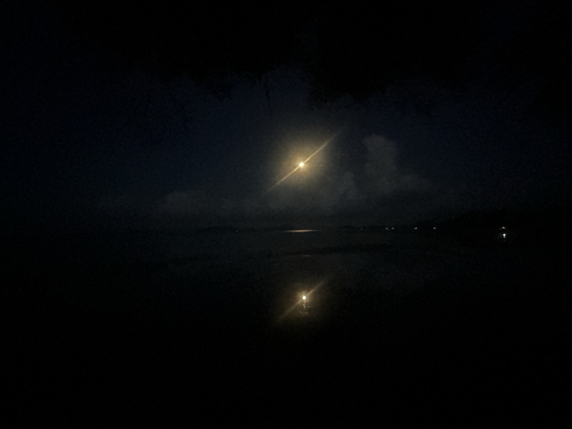 The image shows a night scene in Ban Bang Khao, Thailand, featuring a body of water with a bright reflection of a light source, possibly the moon, in the center. The upper portion of the image shows the light source in the night sky partially obscured by cloud cover. In the distance, faint lights are visible along the shoreline. The foreground is mostly dark, obscuring details.