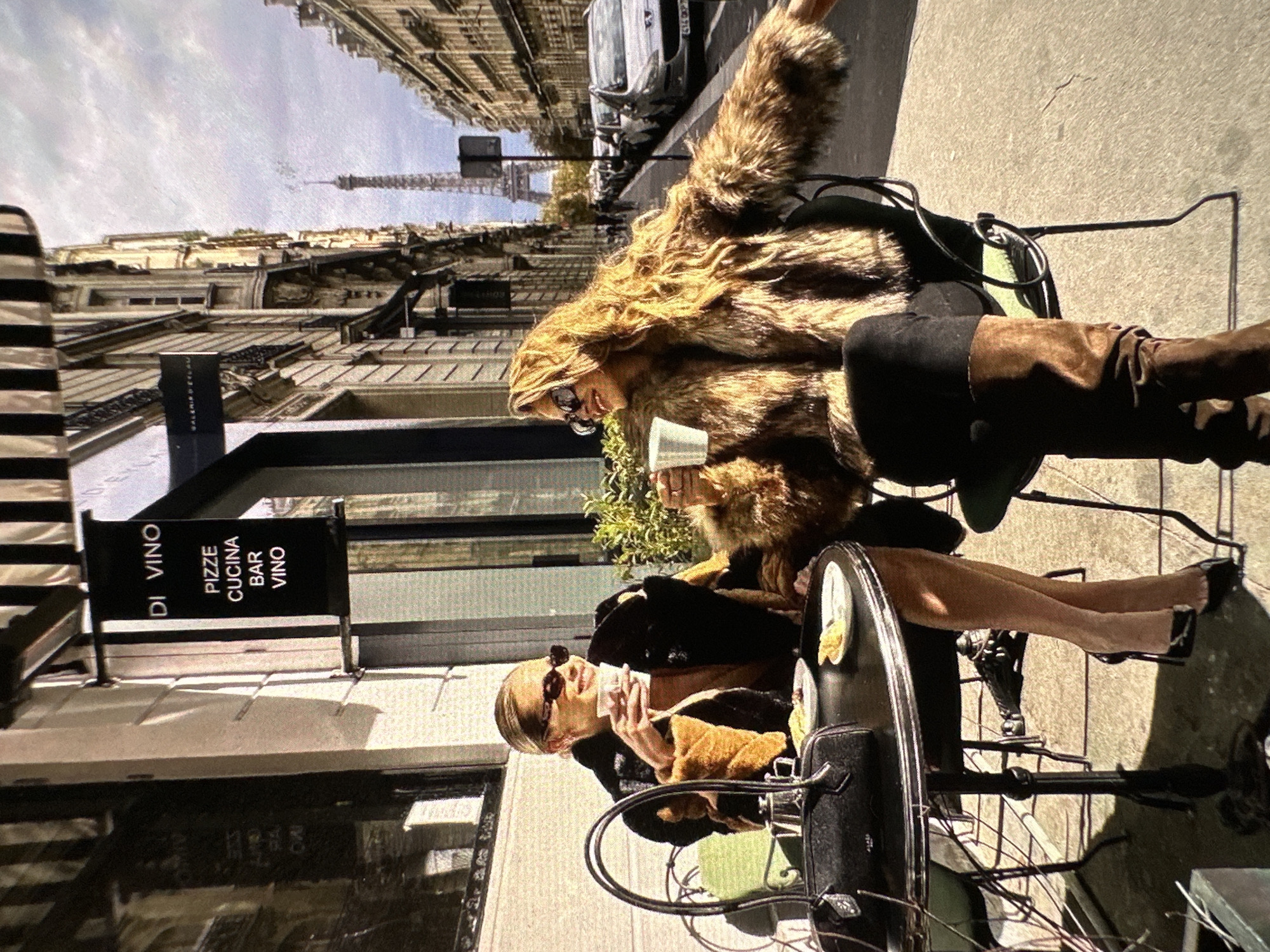 Two women are sitting at a table outside a cafe. The woman on the left is wearing a tan and black jacket. The woman on the right is wearing a large fur coat, a black dress, and brown boots. A sign for "DI VINO PIZZE CUCINA BAR VINO" is visible behind them. The Eiffel Tower is visible in the distance. The location is not Yerevan, Armenia.