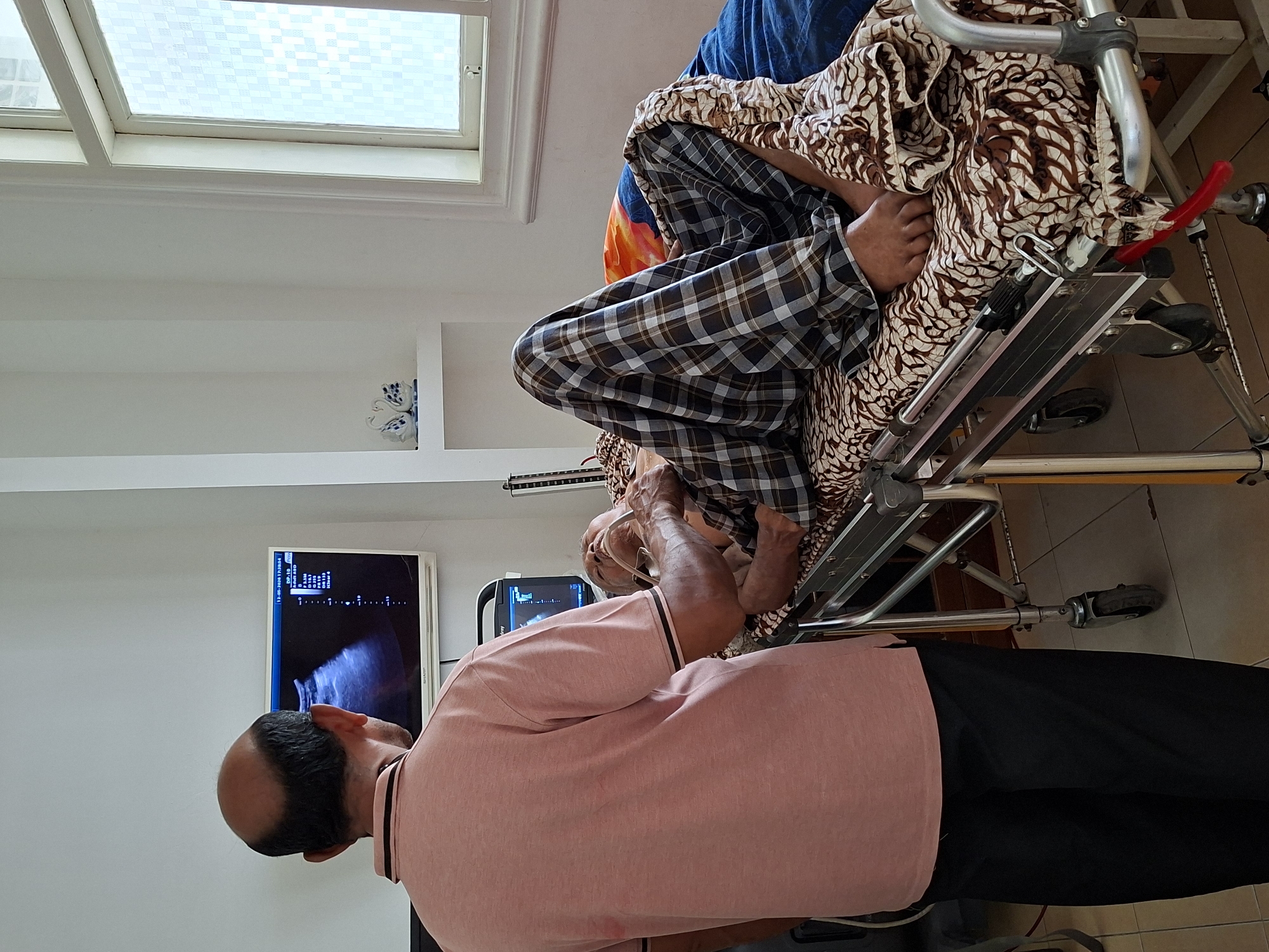 The image shows a man in a pink shirt and black pants looking at a monitor displaying an ultrasound image. A person covered in blankets is lying on a medical stretcher bed. The scene appears to be indoors, possibly in a medical facility. The location is claimed to be in the city of Uleereuleung, Indonesia.