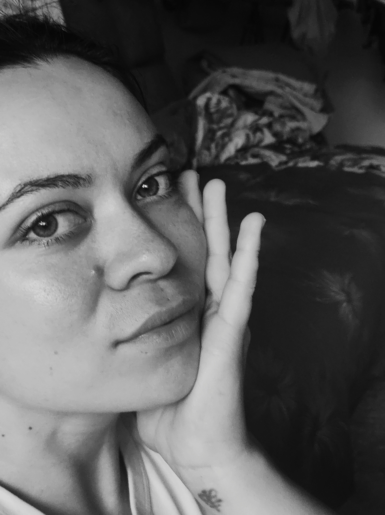 A close-up, black and white portrait captures a woman with her face tilted slightly to the left. Her right hand is positioned with fingers pointing upwards and resting against her cheek. She has dark eyebrows and eyes, a prominent nose, and full lips. A small stud adorns her nostril. A small tattoo is visible on her forearm. The background shows indistinct shapes and textures that suggest bedding. The location in the city of Chaikovsky, Russia is not discernible in the image.