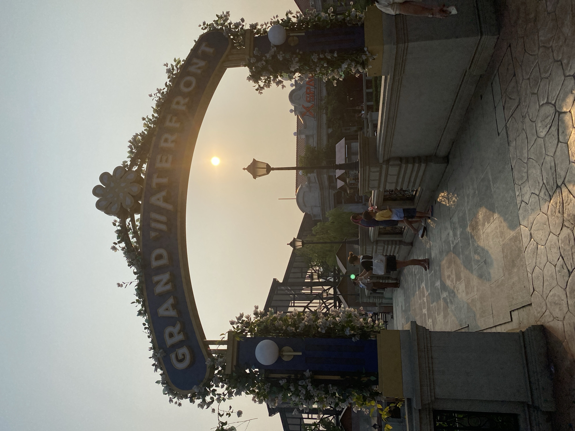 The image depicts a street scene during what appears to be either sunrise or sunset, given the warm, hazy sunlight. A large, arched gateway with the text "GRAND WATERFRONT" prominently displayed is a central feature. The arch is adorned with artificial flowers and greenery, as well as decorative lights.
Several people are visible in the foreground and midground, appearing to be strolling or gathering. Their attire suggests a casual setting. The ground is paved with large, hexagonal stone tiles, and there are ornate lampposts and architectural elements reminiscent of a promenade or a themed area. In the background, buildings with red tiled roofs and some signage can be observed. The overall atmosphere suggests a public space, possibly a tourist attraction or a recreational area. The time of day is indicated by the low angle of the sun and the warm, diffused light, casting long shadows. The weather appears to be clear, though the haze might suggest a slightly humid environment. The location context provided indicates East Jakarta, Indonesia.