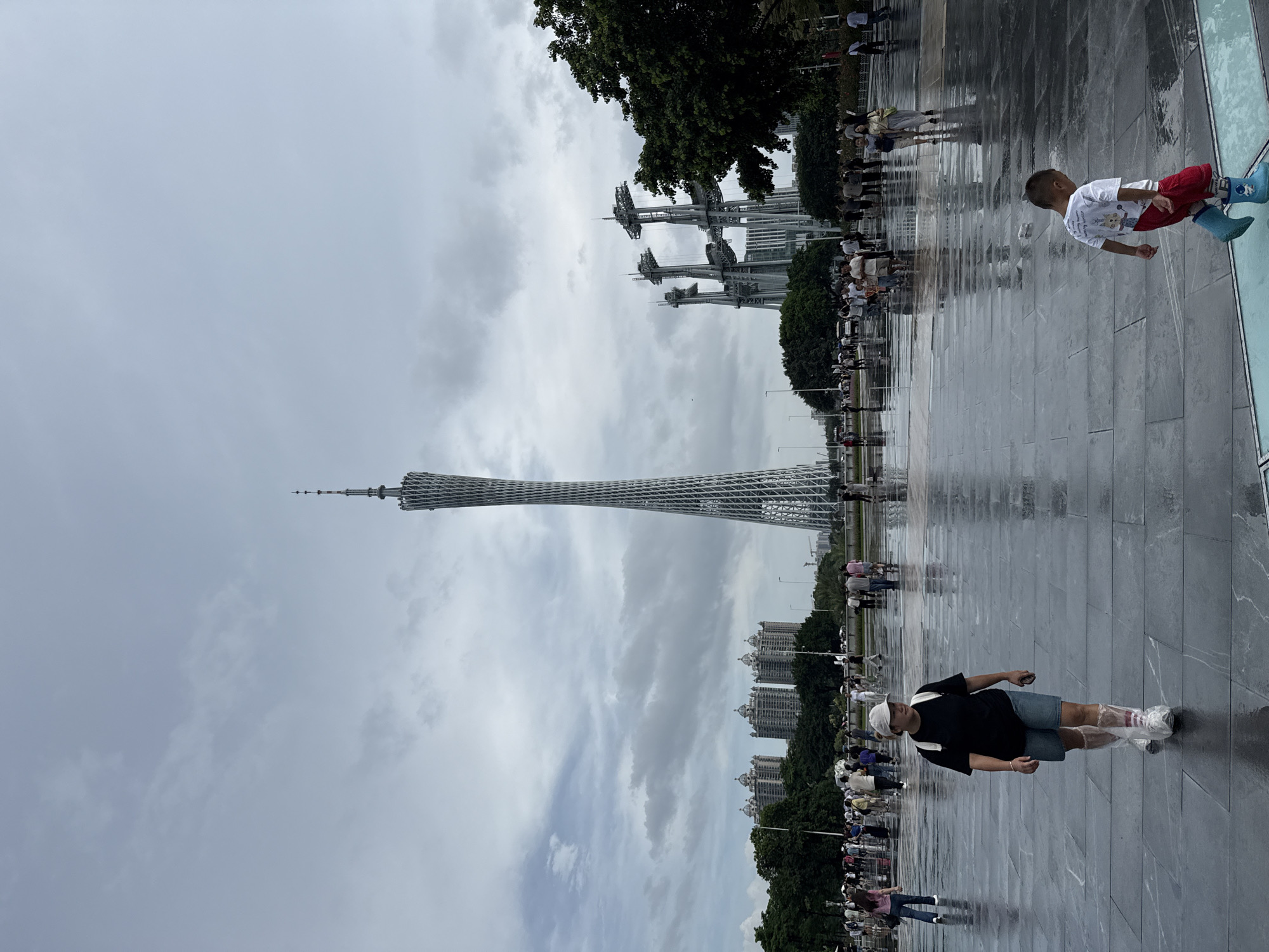The image depicts a public square in Tianhe District, China, featuring the Canton Tower as a prominent landmark. The ground is wet, reflecting the overcast sky and the surrounding structures. Several people are visible walking across the square. In the foreground, a woman wearing a dark t-shirt, denim shorts, and clear plastic shoe covers is walking towards the viewer. To the right, a young boy in a white t-shirt and red shorts, wearing blue rubber boots, walks away from the viewer. In the background, the Canton Tower stands tall, a modern lattice-work structure. To the right of the Canton Tower, a cluster of metallic structures with suspended elements are visible. Several multi-story buildings and trees line the perimeter of the square, with a crowd of people gathered further in the background. The overall atmosphere suggests a humid or recently rained-on day.