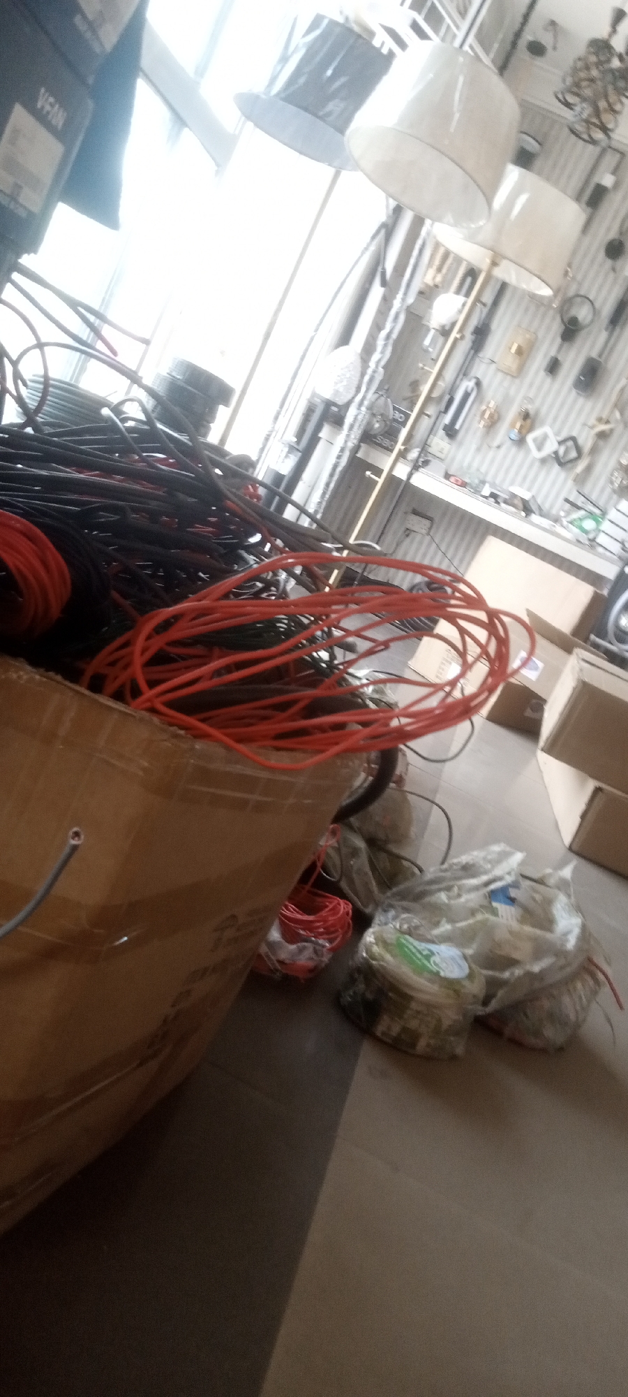The image depicts a cardboard box filled with bundled red and black electrical wires. Adjacent to the box are several plastic bags containing coiled wire. In the background, a window provides bright daylight, revealing a partial view of lamps and other items suggestive of a lighting or electrical supplies store. The image was taken indoors, likely within a shop in Sardauna, Nigeria. No people are visible.