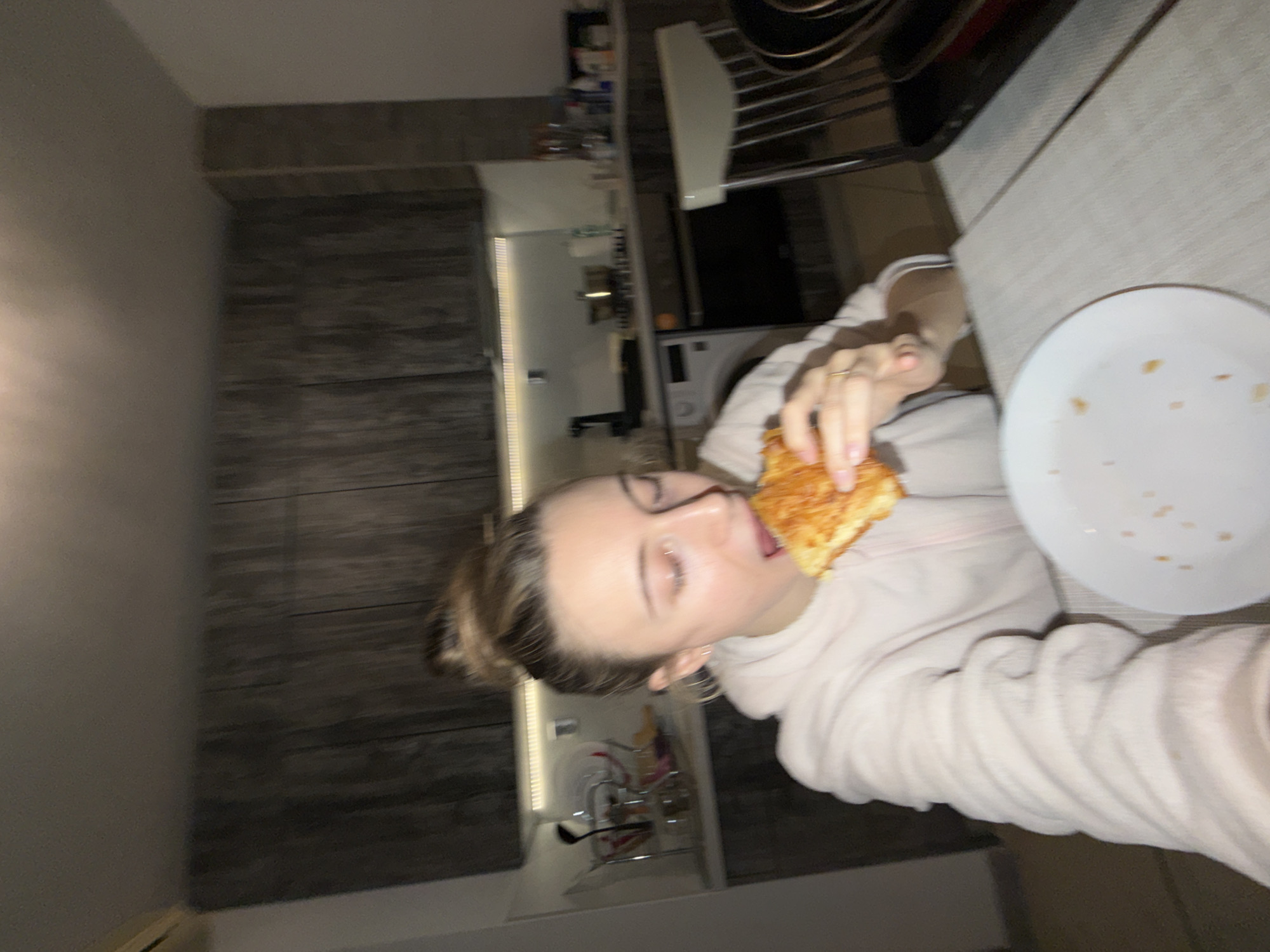 The image presents a scene inside a residential kitchen and dining area in Plovdiv, Bulgaria. A single female subject, with light skin and light brown hair tied in a bun, is depicted in the foreground, actively consuming a piece of what appears to be a savory pastry or cheesy bread. Her mouth is open as she bites into the food, which she holds with both hands. She wears a light-colored, possibly off-white or light pink, fuzzy hooded garment. A simple ring is visible on her left ring finger.
In front of the subject, a white plate containing scattered orange-brown crumbs rests on a light-colored table surface. To the right of the subject, a segment of a light-colored chair with vertical back slats is visible, partially obscuring dark, stacked kitchenware.
The background reveals a modern kitchen with dark grey or brown, wood-grain patterned upper and lower cabinets. A white countertop is present, illuminated by an under-cabinet LED strip light. Various kitchen items are arranged on the countertop, including a dish drying rack holding white dishes and dark utensils, pots on a stovetop, and assorted bottles or containers. A white washing machine or dishwasher is situated next to a dark built-in oven. The scene is illuminated by artificial indoor lighting, and the image exhibits slight motion blur, particularly around the subject's face.