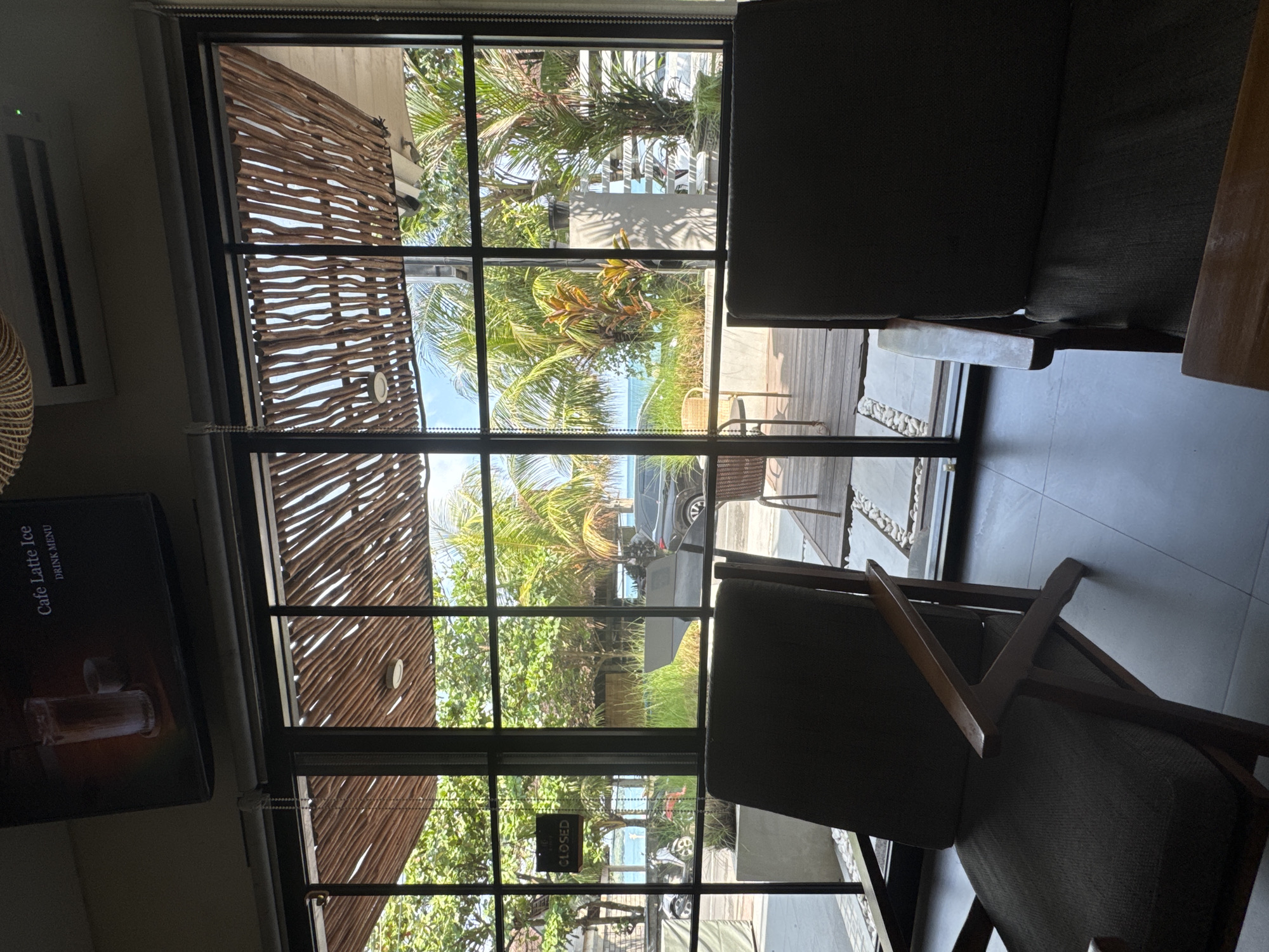 View from an indoor seating area of a cafe in Kuta, Indonesia, looking out through a multi-paned window. The foreground includes two dark chairs, one positioned to the right and one towards the bottom center of the frame. Outside the window, there is a view of a beach with palm trees and other vegetation. A building with a wooden slat exterior is visible to the left of the window. A sign reading "CLOSED" is visible in the window, near the bottom center. A partially obscured menu with the words "Cafe Latte Ice" is seen on the left wall inside the cafe. A grey vehicle is also visible through the window.