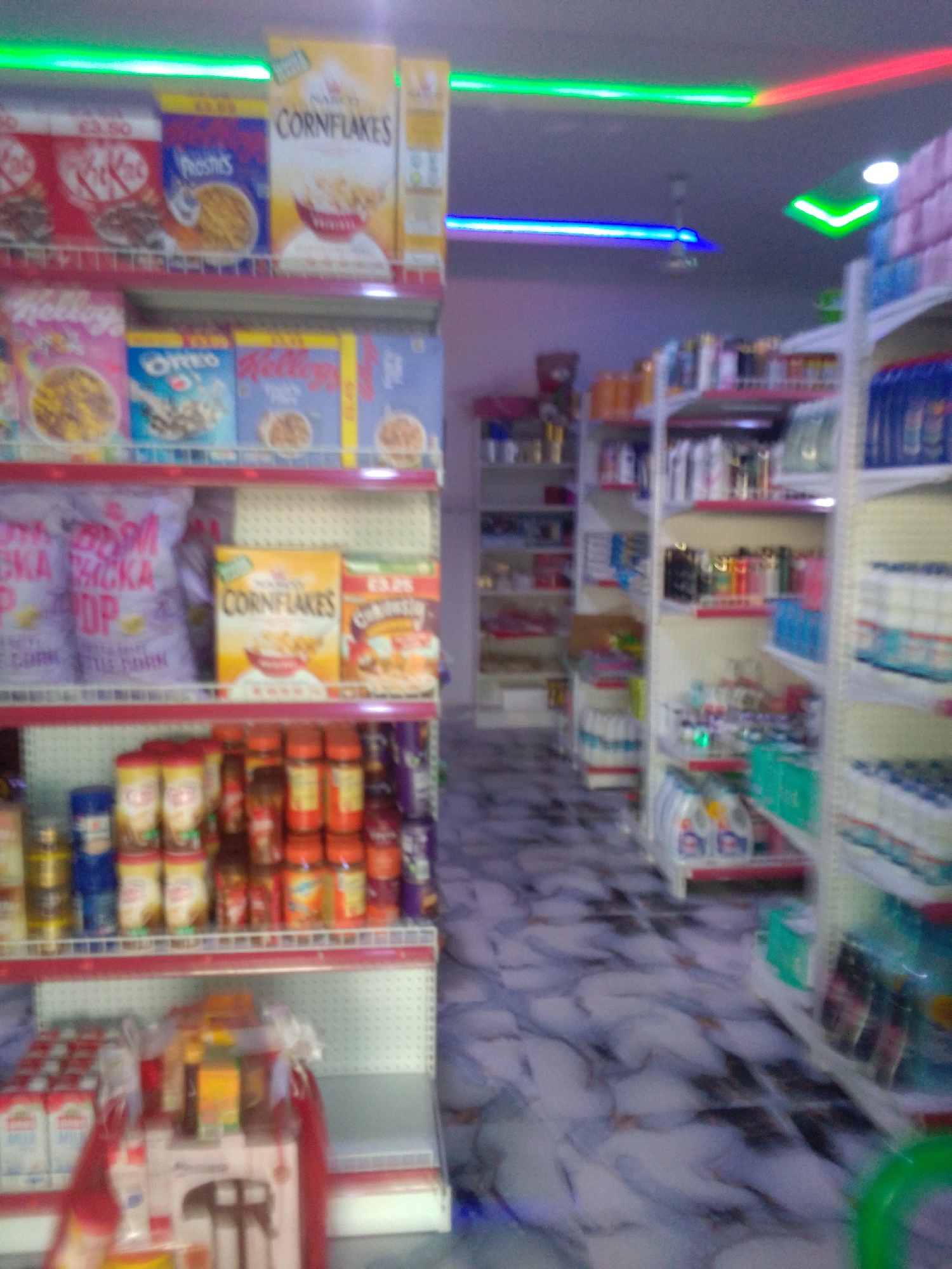 This is a medium shot, eye-level, indoor photograph of a store aisle, likely a convenience store or supermarket, in Howayi, Nigeria. The aisle is lined with shelves stocked with various grocery items, predominantly cereals and packaged foods.
On the top shelf, visible on the left, are boxes of "KitKat" and "Kellogg's Frosties" cereals. To the right of this, a prominent box of "Kellogg's Cornflakes" is displayed. Further to the right, partially obscured, are more cereal boxes.
The middle shelf features larger bags of what appears to be snack food, with the words "CHICKA POP" visible on one. Next to these are more boxes of "Kellogg's Cornflakes" and another cereal box with a price of "£3.25".
The bottom shelf displays an assortment of jars and bottles, many with orange and yellow labels, possibly condiments or spices. These are arranged in rows. On the far right of this shelf, some dark purple packaging is visible.
In the foreground, on a lower shelf or display stand, are stacks of rectangular, white-packaged items, likely milk cartons or juice boxes, some with red ribbon accents. A display with what looks like a food mixer appliance is also present.
The store's interior features bright, colorful LED lighting strips along the ceiling, in green, blue, and red. The floor has a patterned tile design. The overall impression is a well-stocked retail environment. No individuals are visible in the frame.