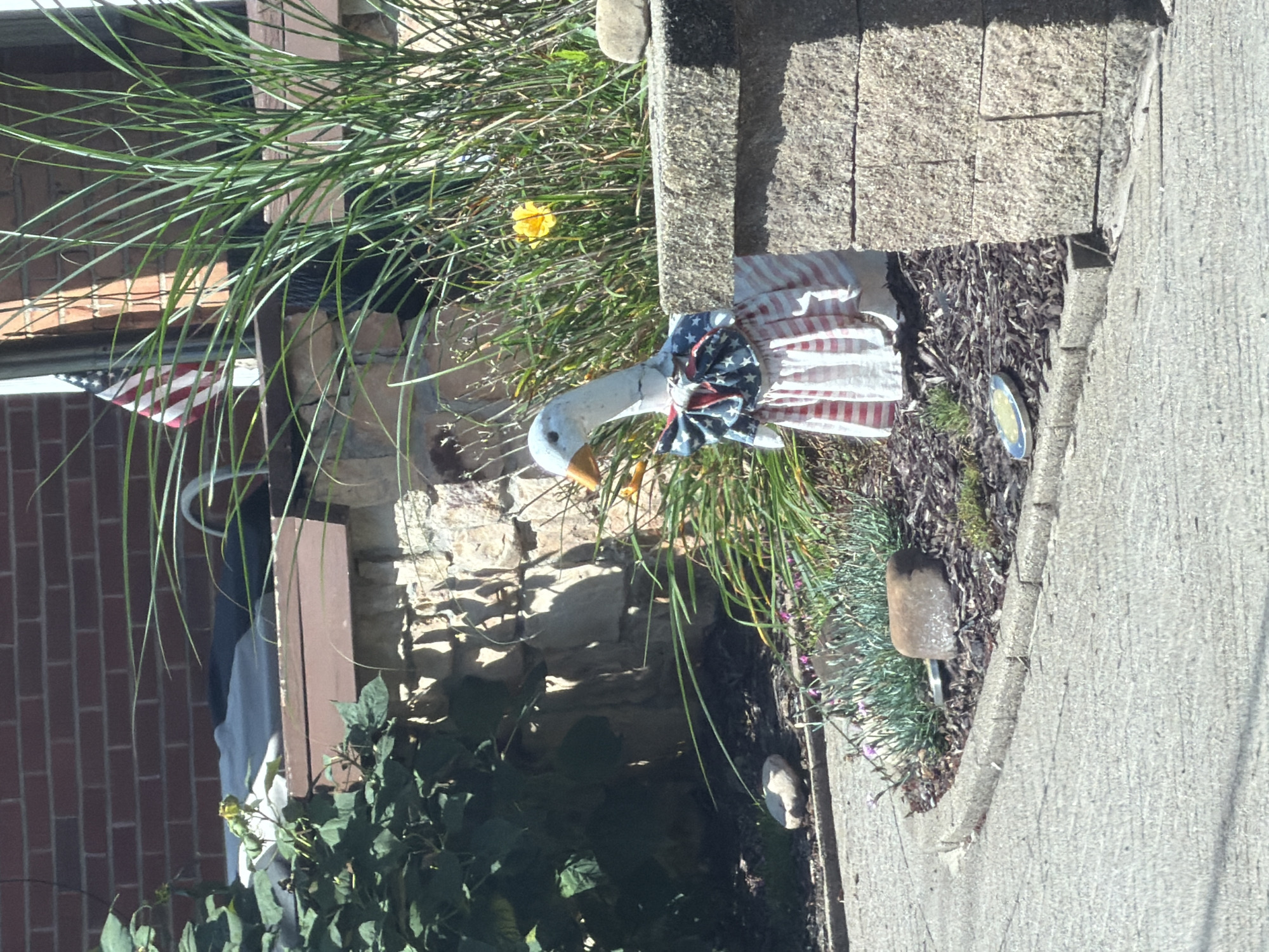 The image shows a decorative goose figurine wearing a patriotic bow tie and dress, placed in a garden area next to a stone wall. Behind the goose, tall green grass and a yellow flower are visible. To the left, a brick wall and some foliage are present. An American flag is partially visible in the background. The ground in front of the goose is paved with concrete. The overall scene appears to be an outdoor residential setting, likely a front yard or garden. The location is identified as Penn Hills, United States of America.