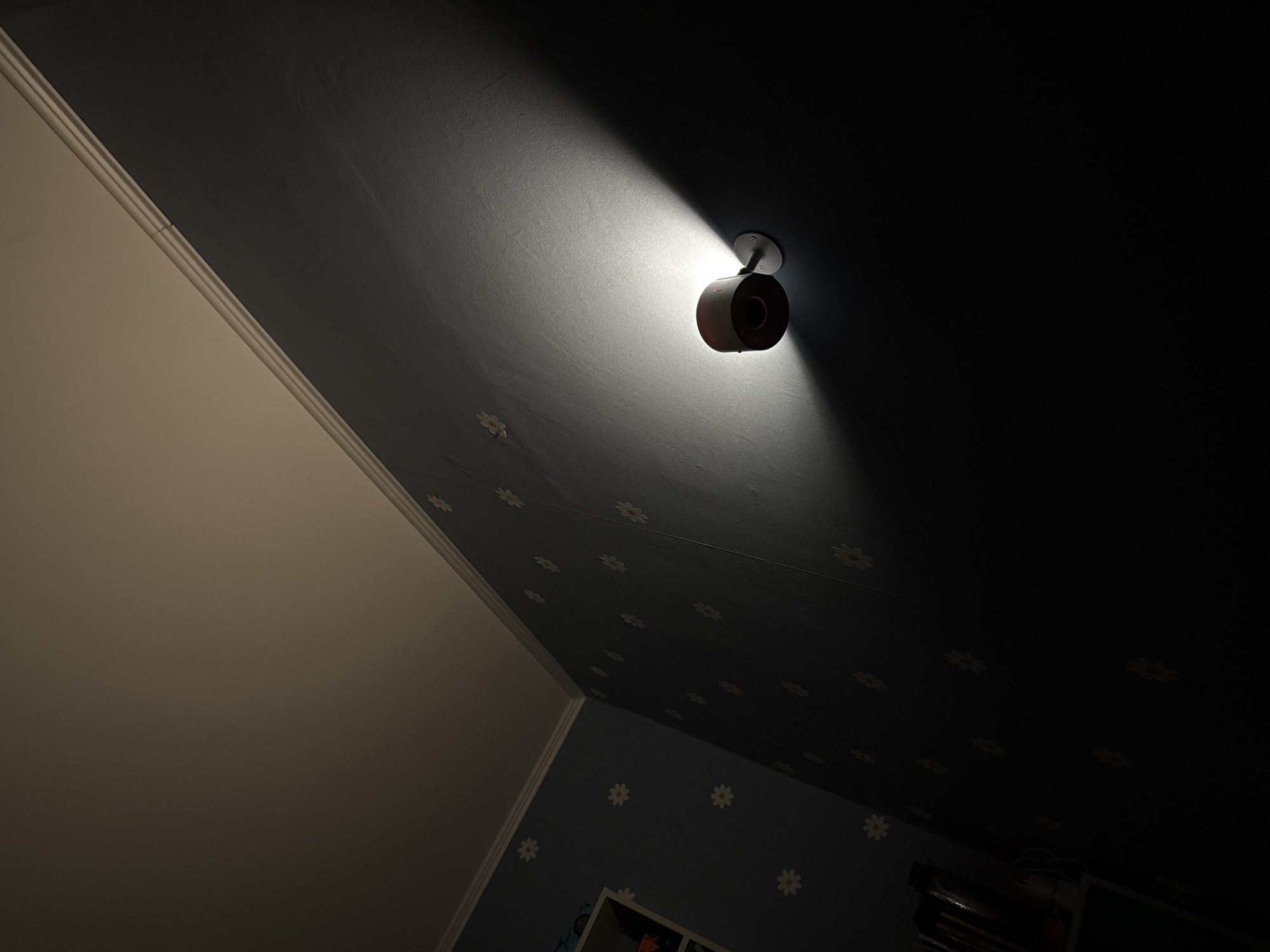 A dim, low-angle interior shot shows a security camera mounted on a blue wall adorned with white daisy patterns. The camera is positioned towards the upper-right of the frame. A focused beam of light emanates from the camera, illuminating the wall directly in front of it. To the left, a beige wall extends into the scene. The lower portion of the frame reveals a glimpse of a wall and the top of furniture, also decorated with white daisy patterns. The location is not identifiable as being in Moscow, Russia.