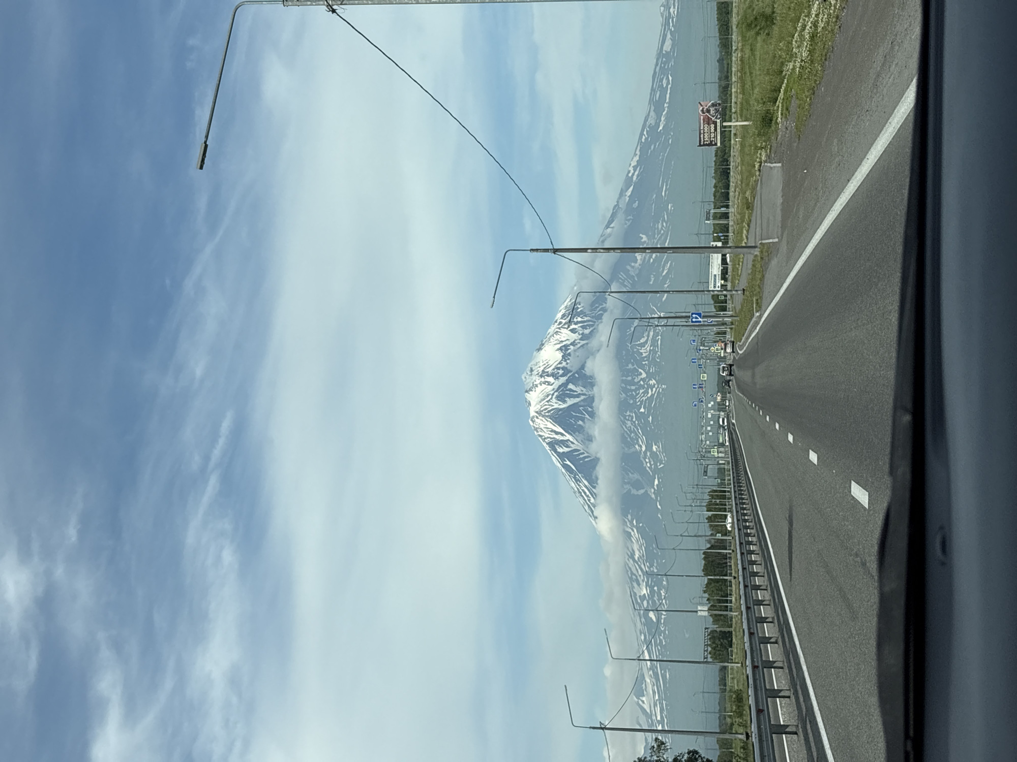 The image depicts a view from a vehicle traveling on a highway towards a snow-capped volcano. The volcano dominates the background, with clouds partially obscuring its lower slopes. The highway is paved with asphalt and has white lane markings. There are streetlights on both sides of the road, and a metal guardrail runs along the left side. In the distance, trees line the horizon, and there are several cars on the highway. A billboard is visible on the right side of the road. The sky is blue with scattered white clouds. The scene appears to be in or near the city of Yelizovo, Russia, given the presence of the prominent volcano, which is likely Mount Avachinsky.