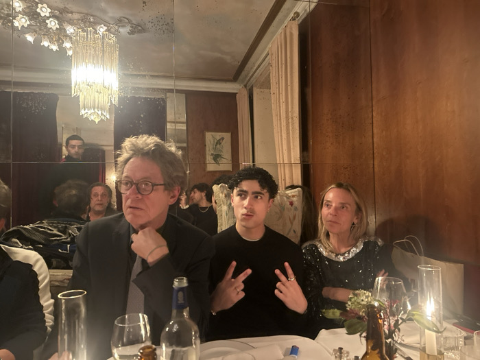 The image portrays an indoor dining scene at a restaurant in Paris, France. Three individuals are seated at a white-clothed table, illuminated by dim ambient lighting and a central candle. On the left, a middle-aged male with wavy brown-grey hair and spectacles, dressed in a dark suit jacket and tie, looks toward the left with his right hand positioned near his chin. Central to the frame, a young male with dark curly hair, wearing a black long-sleeved shirt, makes a "peace" sign with both hands while looking directly forward. To his right, a middle-aged female with pulled-back blonde hair, wearing a black top adorned with silver sequins, sits with her arms crossed, looking slightly to her right.
The table is set with clear drinking glasses, a dark-colored bottle, a small floral arrangement featuring green leaves and dark red blossoms in a brown vase, and a lit pillar candle contained within a clear glass holder. The background consists of aged, segmented mirrors covering the walls, which reflect a prominent multi-tiered crystal chandelier, a second chandelier with floral glass elements, light-toned curtains, wooden wall panels, and indistinct figures of other patrons. The overall environment suggests an intimate, upscale evening gathering.