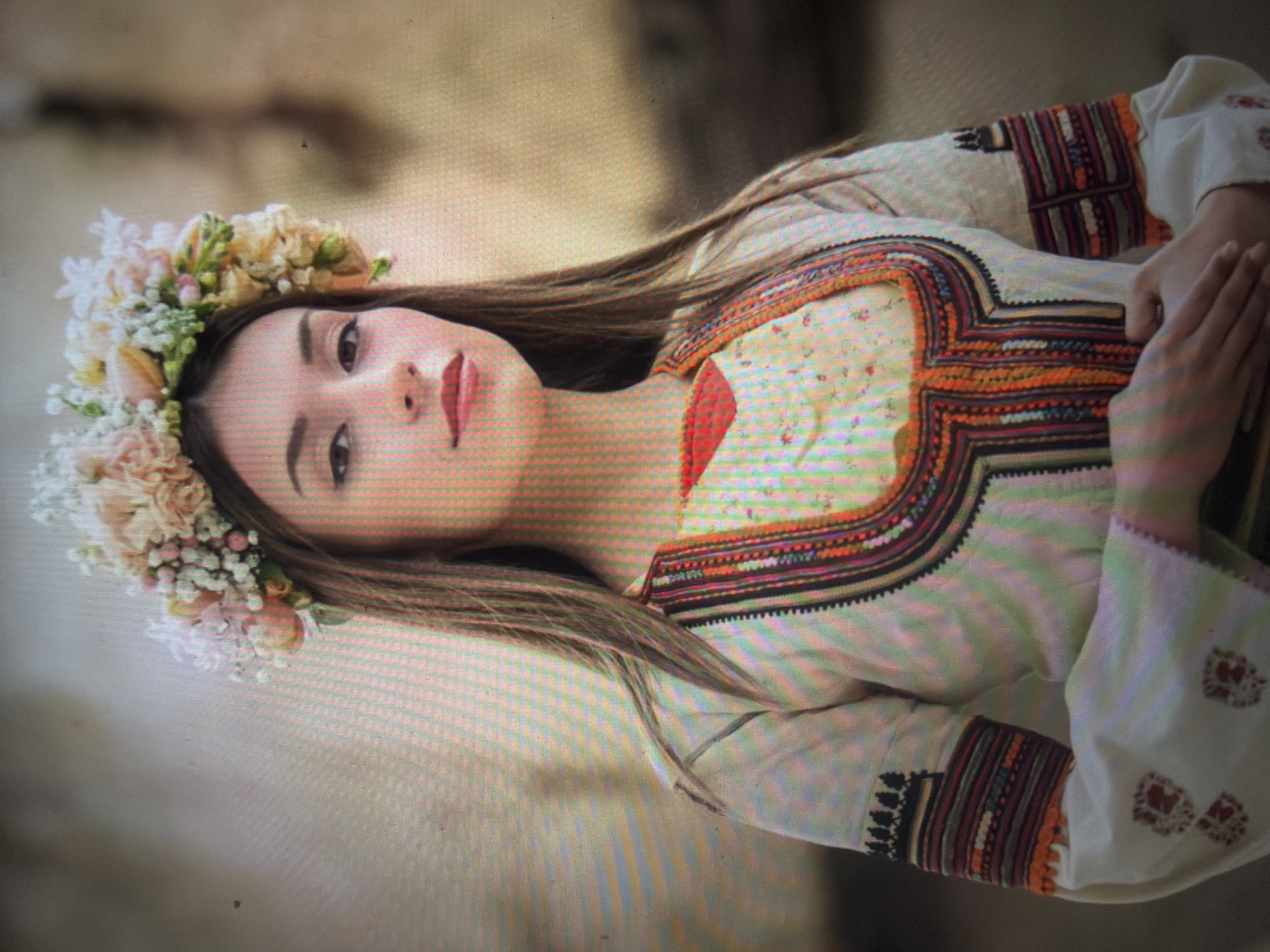 A young woman is depicted in a waist-up portrait, centered and facing directly forward. She appears to be in her late teens or early twenties, with long, straight dark brown hair falling over her shoulders. She wears a floral crown adorned with light pink, white, cream, and yellow flowers, interspersed with green foliage. Her dark eyes are open, and her lips are colored with a pink or light red lipstick, maintaining a neutral and direct gaze towards the viewer. She is dressed in a traditional garment, specifically a light-colored blouse featuring elaborate, multi-colored embroidery. The embroidery around the neckline forms a distinct U-shaped pattern in shades of red, orange, black, and possibly dark blue or green. Similar intricate embroidery is visible on the upper arms and cuffs of the sleeves, with the cuffs displaying prominent red-patterned stitching. Her hands are clasped together in front of her lower torso. The background is softly blurred and out of focus, suggesting an outdoor setting under natural, diffused lighting, characterized by soft, earthy tones. This scene is located in the city of Shumen, Bulgaria.