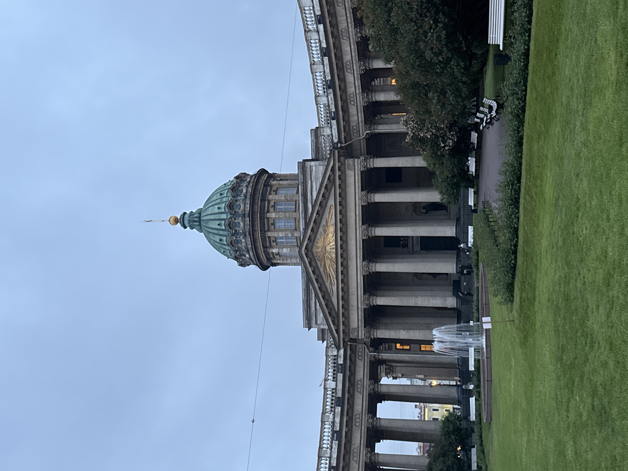 The image depicts Kazan Cathedral in Saint Petersburg, Russia, under an overcast sky. A prominent green dome with a golden spire tops the neoclassical building. The cathedral features a semicircular colonnade with numerous Corinthian columns and a balustrade. In the foreground, a well-maintained green lawn surrounds a circular fountain with water jets. Several white park benches are visible in the background, arranged along a path. A few individuals are present, some walking near the colonnade and others seated on benches. Lush green trees and bushes add to the landscape. The overall scene conveys a sense of grandeur and tranquility.