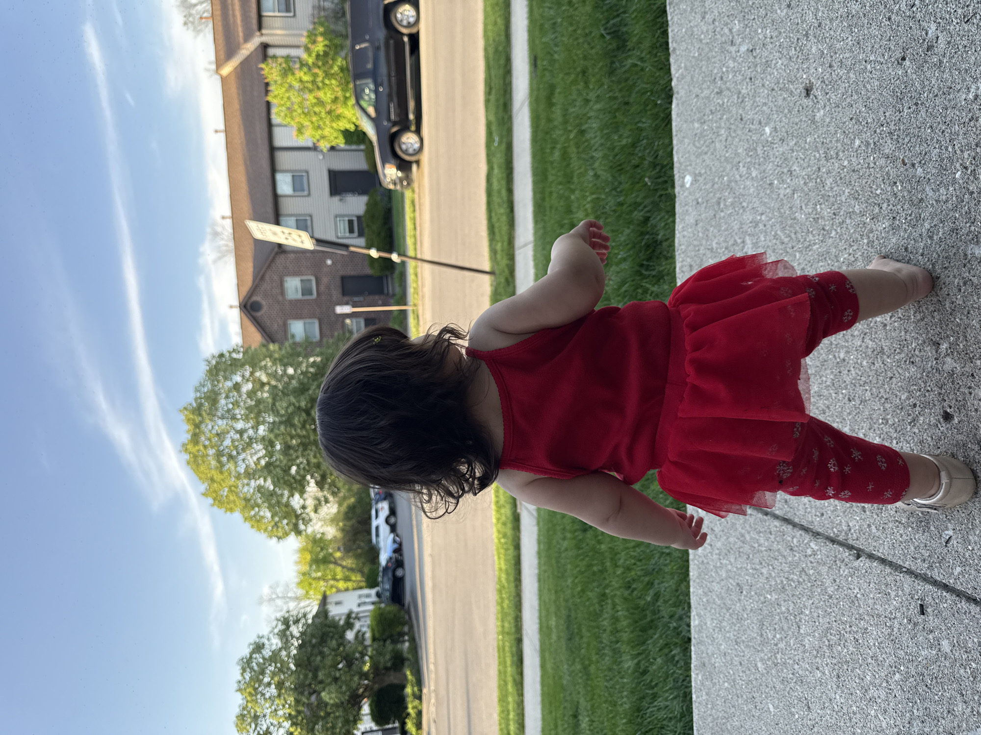 A toddler wearing a red dress and patterned leggings with gold shoes is standing on a concrete sidewalk. The background features a residential street in the city of Columbus, United States of America, with green grass, trees, houses, cars, and a street sign visible. The sky is blue with light clouds.
