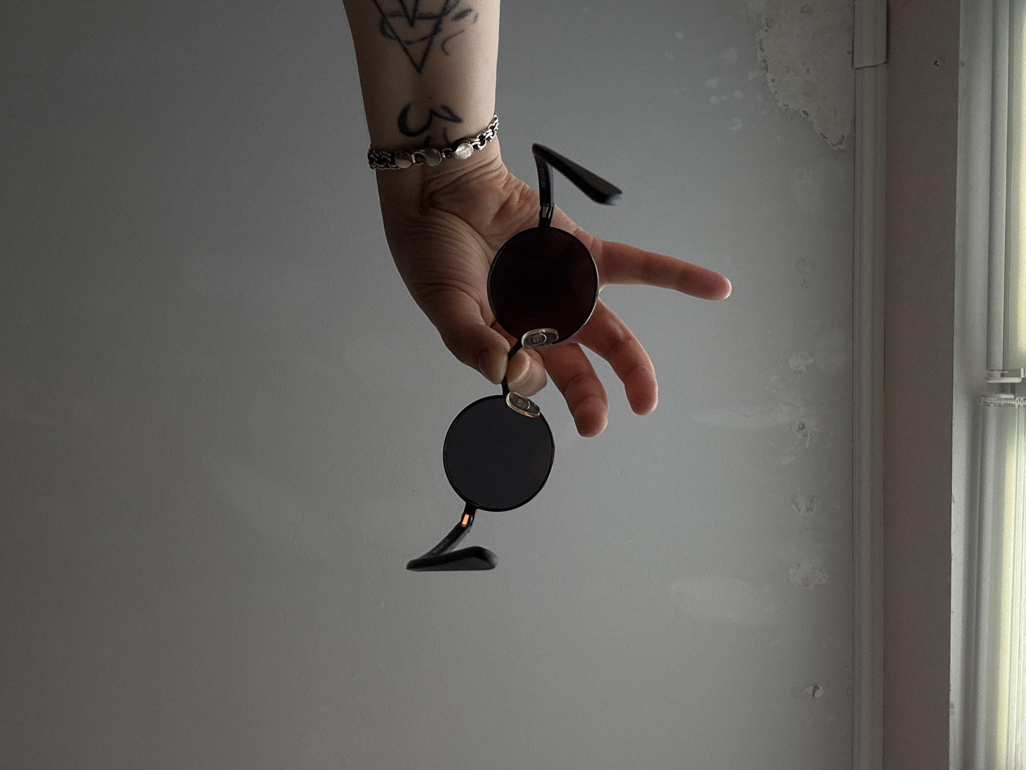 An arm with tattoos and a bracelet holds a pair of round, dark-lensed sunglasses against a gray, textured ceiling. The sunglasses are dark and have thin frames, with one lens facing the camera and the other angled away. The arm is positioned in the upper right portion of the frame. The ceiling appears to have water stains and imperfections. Below the ceiling is a white ledge or molding, and further down, a window with blinds is visible. The overall scene suggests an indoor environment, possibly a room with a damaged ceiling, with no specific identifiable landmarks to place it within Saint Petersburg, Russia.