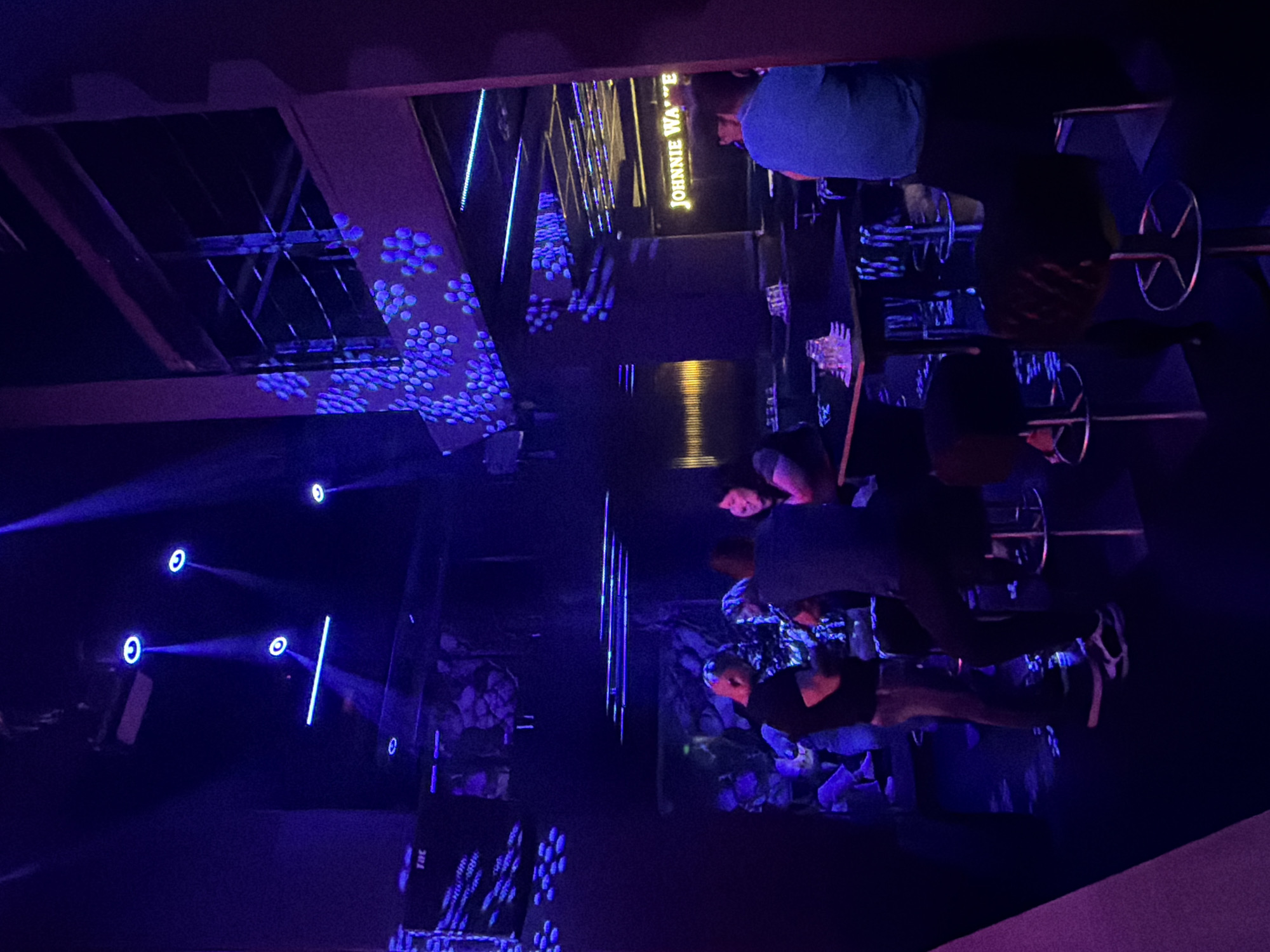 The image depicts a dimly lit interior, likely a bar or nightclub, with extensive use of blue and purple lighting. In the foreground, a person in a blue shirt is seated on a stool, facing away from the camera. To their left, a small cluster of people are gathered around a table. Further left, two individuals stand near a bar, one in a dark t-shirt and grey pants, the other in a dark t-shirt and dark pants. Above and behind them, decorative blue lights illuminate the ceiling and walls, which also feature a pattern resembling blue flowers. In the background, a neon sign reading "JOHNNIE WA." is visible. The overall atmosphere suggests a social gathering in an entertainment venue. No specific location within Veliko Tarnovo, Bulgaria can be identified from the image.