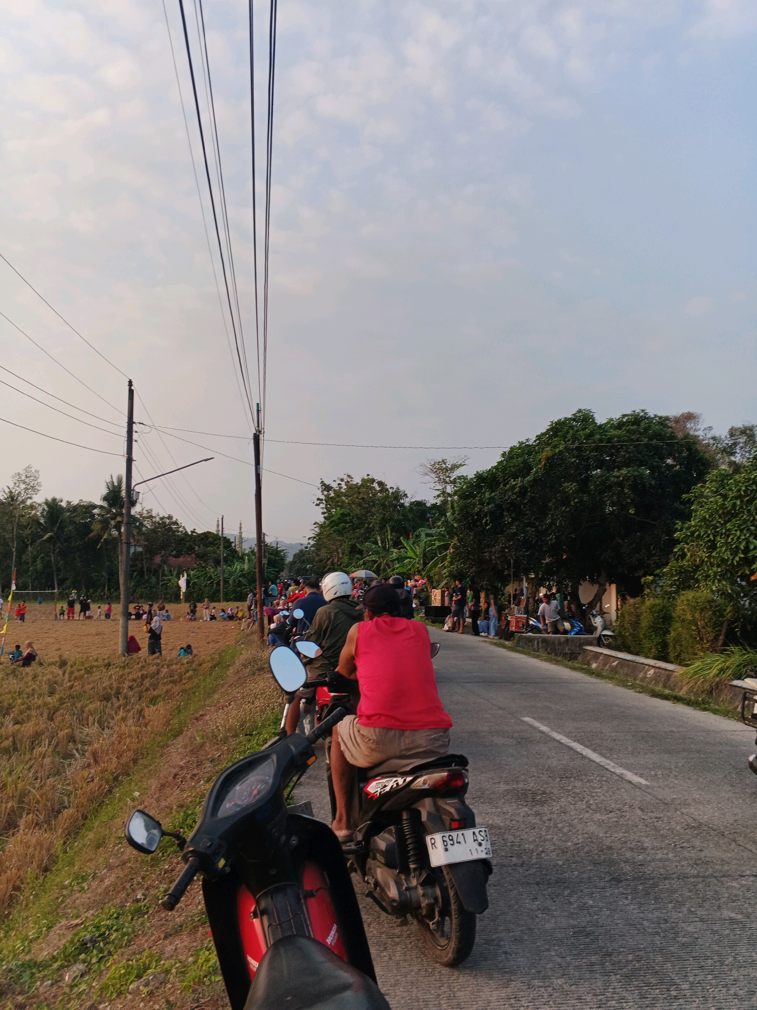 In Kuripan, Indonesia, a street scene unfolds with a focus on a line of motorbikes. The foreground is dominated by the front of a black and red motorbike, its handlebars and mirrors visible. Behind it, a rider in a bright red t-shirt and brown shorts is seated on a motorbike, the rear of which, with its license plate "R 6941 AS", is clearly depicted. Another motorbike with a white helmet rider is positioned slightly ahead. The road stretches ahead, bordered by a grassy verge on the left and a low stone wall on the right. Beyond the verge, a field of dry, golden grass is dotted with numerous people, some sitting, others standing, and a few children playing. Power lines crisscross the sky overhead, supported by utility poles. In the distance, lush green trees form a backdrop, with glimpses of buildings and what appears to be a mosque or similar structure. The sky is a soft, overcast blue, suggesting it is late afternoon or early evening.