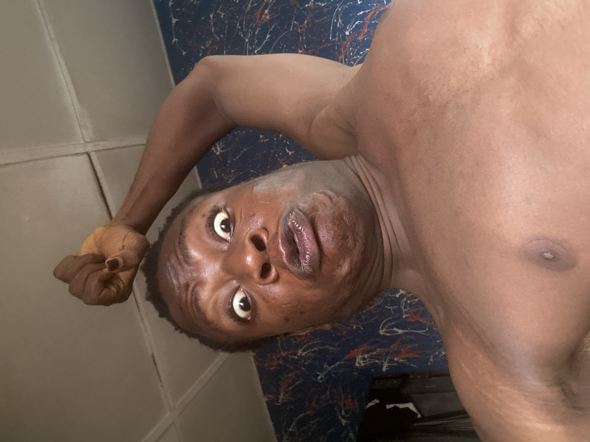 The image shows a person with dark skin and short, dark hair. The person's face is close to the camera and their expression is one of surprise or shock. Their eyes are wide open, and their mouth is slightly open. The person is shirtless, revealing a portion of their chest and shoulder. The background appears to be a room with textured surfaces.