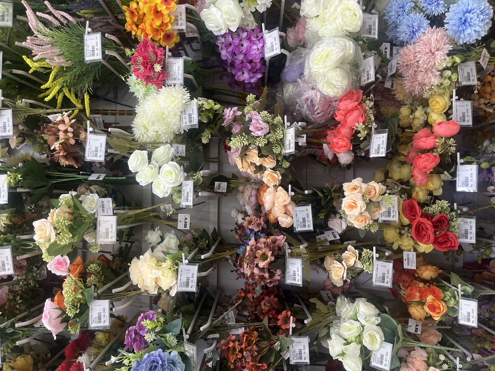 The image displays a vibrant, overhead view of numerous artificial flowers arranged vertically on display hooks, likely in a retail store setting in West Jakarta, Indonesia. The scene is dominated by a wide assortment of synthetic floral bouquets and individual stems, presented for sale.
The artificial flowers come in a rich palette of colors, including various shades of pink, yellow, orange, red, white, cream, purple, and blue, complemented by green foliage. Different floral types are visible, resembling roses, peonies, and other decorative blossoms, creating a dense and colorful tapestry across the frame. Some flower bundles are partially encased in clear plastic packaging.
Each bunch of flowers is accompanied by a white price tag, indicating prices in Indonesian Rupiah (Rp). Notable prices include Rp 14,000, Rp 18,500, Rp 16,500, Rp 19,500, Rp 15,500, Rp 23,500, Rp 17,500, Rp 30,000, Rp 27,000, Rp 13,500, and Rp 16,000. The tags also feature product names such as "ARTIFICIAL FLOWER SH", "ARTIFICIAL ROSE SH 3", and "ARTIFICIAL PEONY 213", along with product codes like "9047772" and "8047709". The "DIY" logo is prominently displayed on many of the tags, suggesting that the store caters to do-it-yourself or craft enthusiasts.
The setting is indoors, with no people visible in the frame. The focus is entirely on the merchandise, presented in an organized and visually appealing manner for customers.