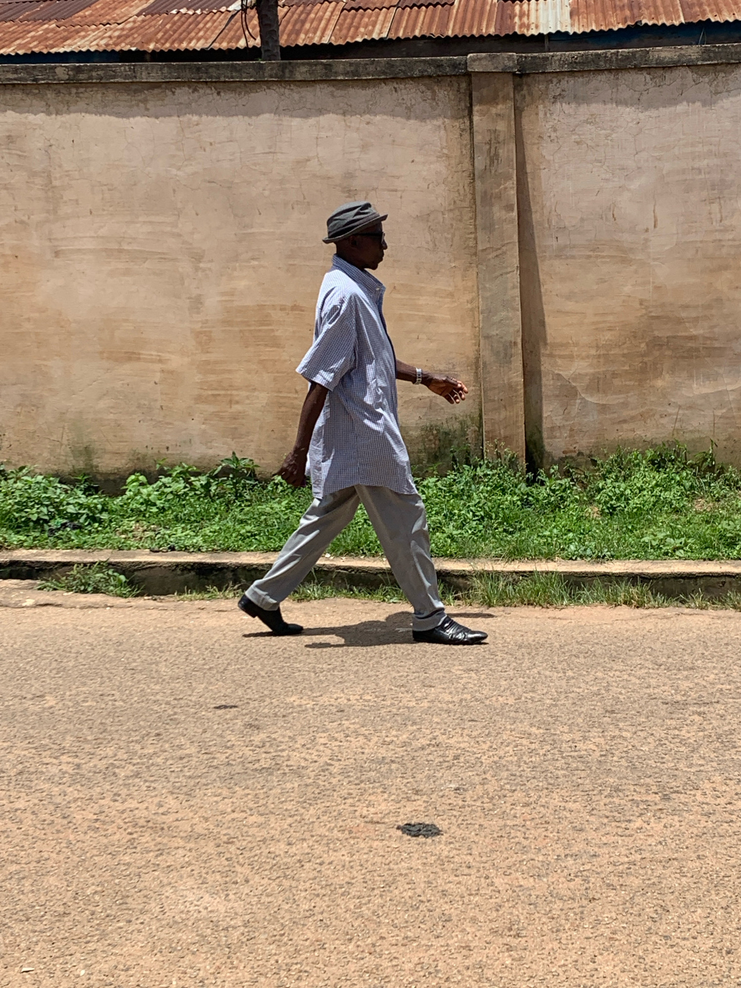 An elderly African man, wearing a grey flat cap, a blue and white checkered shirt, and grey trousers, walks on a paved road. He is positioned in the middle right of the frame and is walking from left to right. The background features a tan-colored wall with some cracks, topped with a rusty corrugated iron roof. Lush green vegetation grows along the base of the wall. The scene is lit by daylight, casting shadows on the road. The overall environment appears to be an outdoor setting in an urban area, potentially Jos, Nigeria, given the architectural style and vegetation.