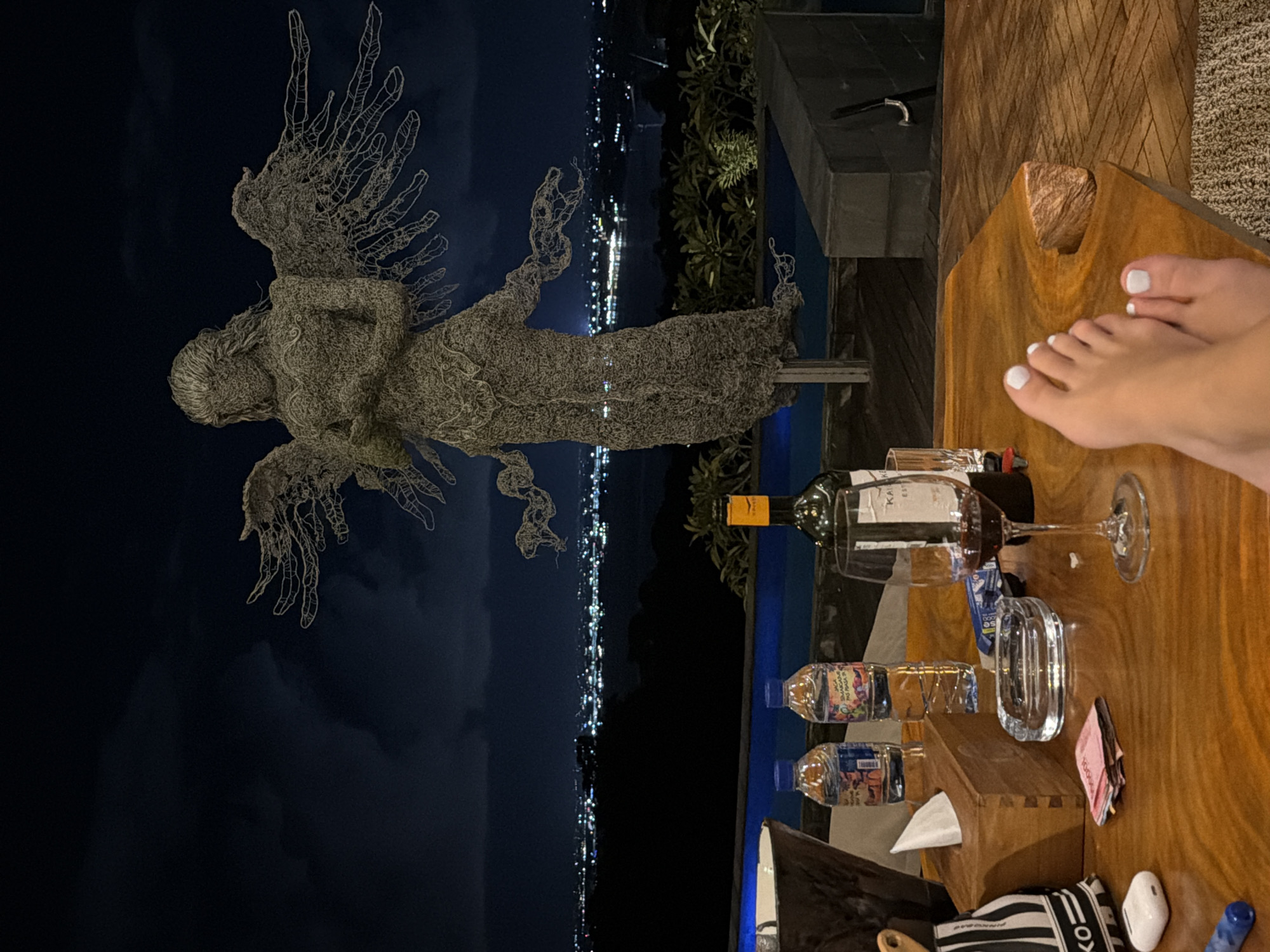 The image shows a nighttime outdoor scene in Kuta Selatan, Indonesia. A metallic sculpture resembling an angel with outstretched wings stands prominently on the left. In the background is a dark sky and a view of city lights stretching along the coastline.
On the right, a wooden table holds a wine bottle, a partially filled wine glass, two bottles of water, an ashtray, tissues in a box, a wad of bills, and other miscellaneous objects. A pair of bare feet, with painted white toenails, rest on the table near the objects. The table suggests a patio setting or outdoor dining area.