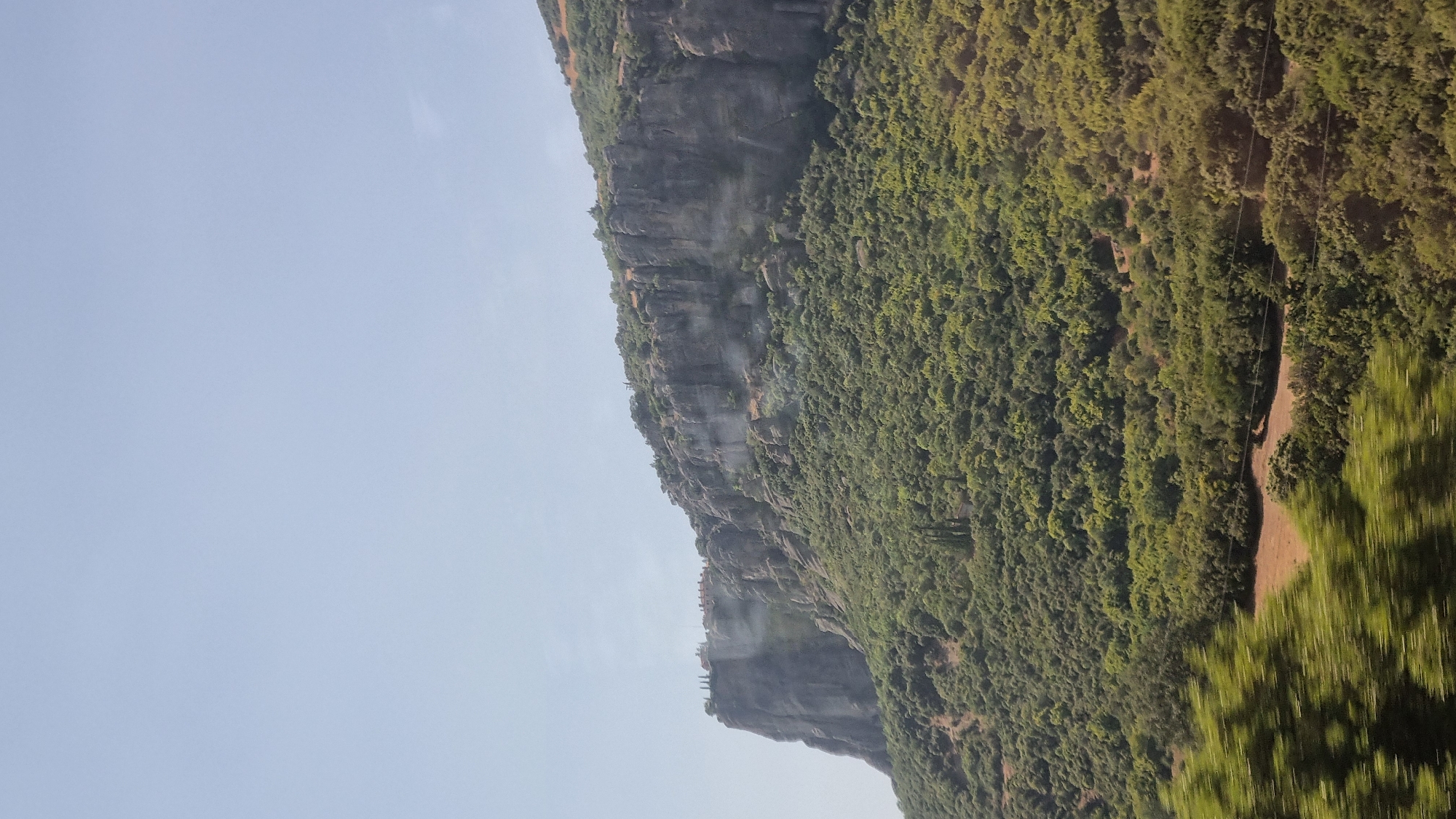 This image depicts a vertical view of a mountainous landscape near the city of Kalambaka, Greece. The upper portion of the image is dominated by a clear, pale blue sky. The middle section features a dramatic, sheer rock face, with a dense, green forest covering its lower slopes and extending into the foreground. Atop the prominent rock formation on the left, several structures, possibly part of a monastery or settlement, are visible. The rocky cliffs exhibit distinct strata and weathered textures. The forested area is lush and varied, with a mix of deciduous and coniferous trees. In the foreground, the greenery appears blurred, suggesting motion, possibly from a vehicle passing by. The overall scene conveys a sense of natural grandeur and historical presence, characteristic of the Meteora region.