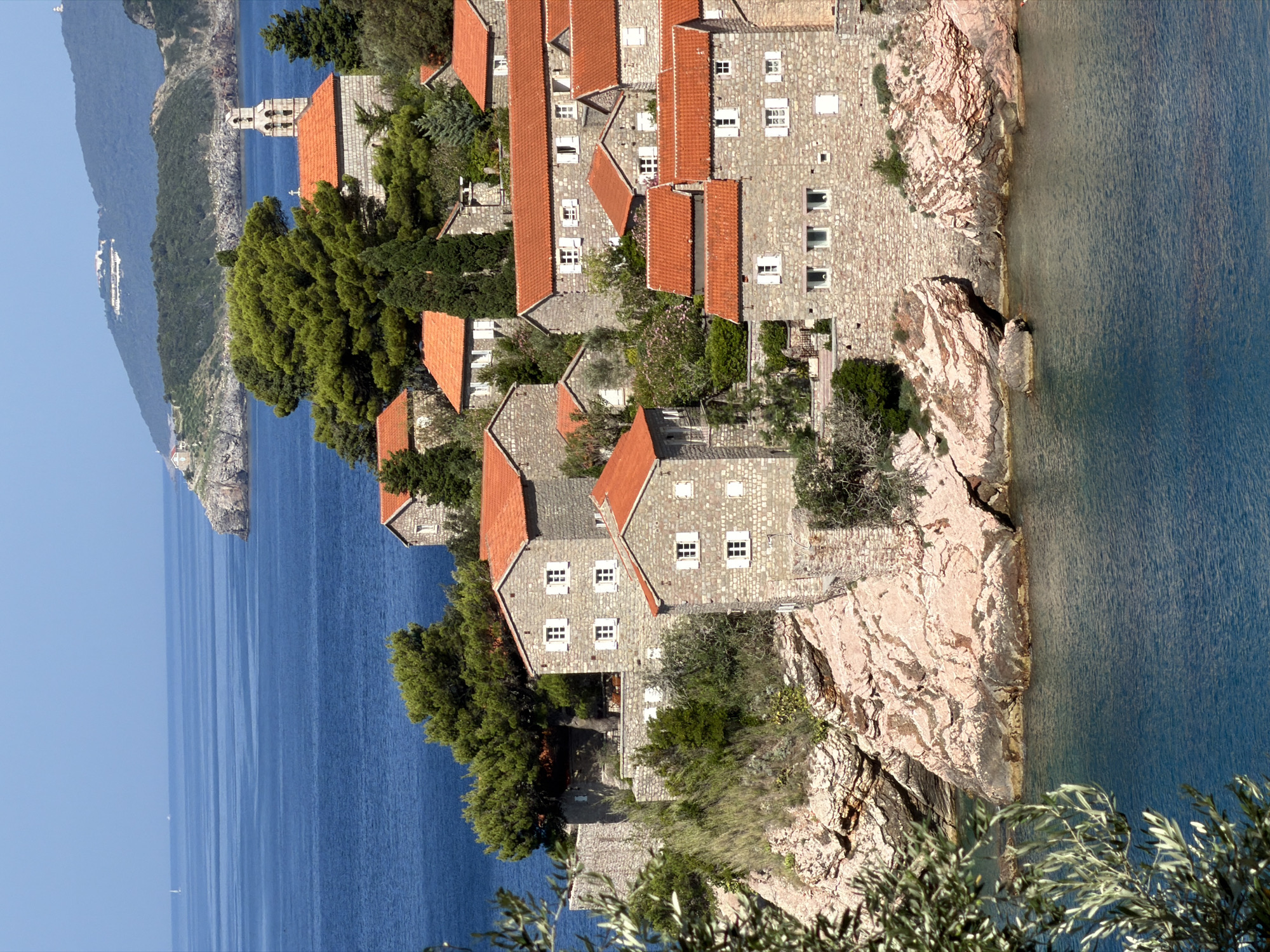The image shows the Sveti Stefan island, a picturesque islet in Montenegro, located off the coast of the Adriatic Sea. The islet is fortified with stone buildings topped with red-tiled roofs, clustered together on a rocky outcrop. Lush green trees and vegetation surround the structures, contrasting with the clear blue of the sea. The architecture suggests a historical settlement, likely a former fishing village, now a resort. The scene is bathed in bright sunlight, casting shadows and highlighting the textures of the stone and the water. The water is calm and a deep blue, with some lighter patches indicating shallower areas near the shore. In the distance, the mainland coastline is visible, with hills covered in green foliage under a clear blue sky. The image captures a serene and idyllic Mediterranean landscape.