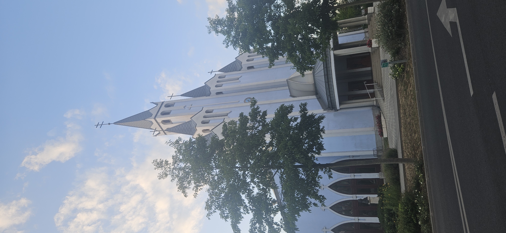 The image shows a light blue church with white trim and dark grey spires, featuring several crosses. The church has arched windows with stained glass and a porticoed entrance. Trees with green foliage frame the building on both sides, and there is a patch of grass and flowerbeds in front. A paved walkway leads to the entrance, and a road with a directional arrow is visible in the foreground. The sky is blue with scattered white clouds. The church is the Holy Spirit Roman Catholic Church located on Fő utca in Hévíz, Hungary. There are no people or animals visible in the image.