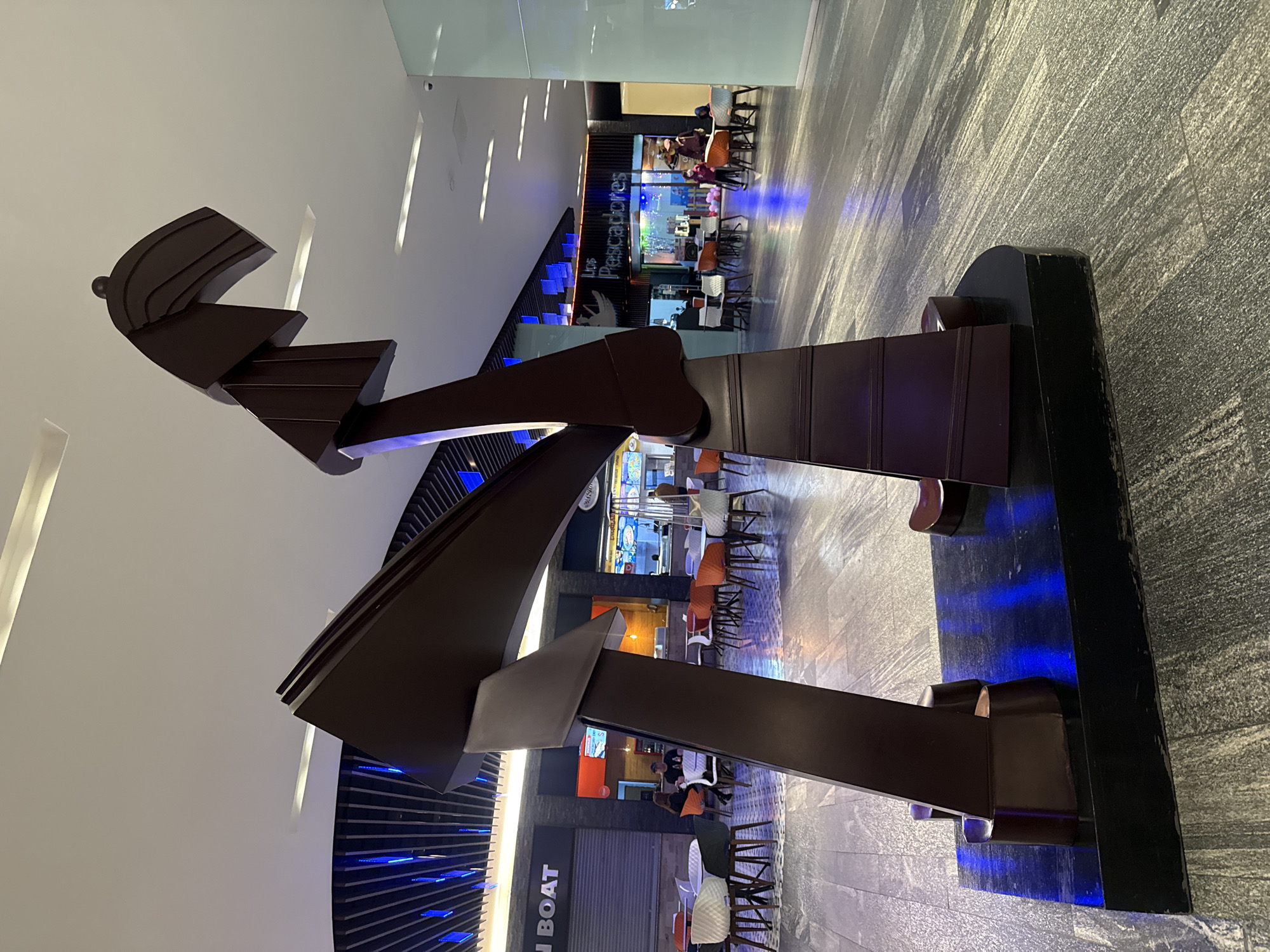 A view within a commercial building, likely a mall or shopping center in Puebla, Mexico. A large, abstract, dark-brown sculpture dominates the foreground, consisting of intersecting beams and planes. The sculpture is mounted on a black rectangular base. The lighting is diffused and somewhat cool, creating a modern ambiance. The floor is a gray, textured material. In the background, partially obscured by the sculpture, are several commercial establishments with tables and chairs, indicating food vendors or restaurants. One establishment has the name "Los Pescadores" visible. There are a few people seated at the tables in the background, and some of them appear to be in transit, suggesting a busy public space. The environment suggests a contemporary indoor setting with a blend of art and commerce.