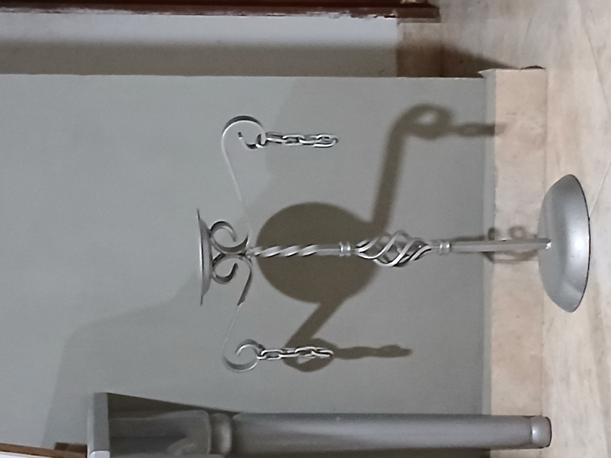 The image depicts a silver metal candelabra with three arms, each supporting a hanging chain. The candelabra is situated on a tiled floor in front of a grey wall. To the left of the candelabra is a portion of a silver-colored column or table leg. The lighting casts shadows of the candelabra and its chains onto the wall. The scene appears to be indoors, possibly in a home or a commercial establishment in Santa Ana, Venezuela. There are no people visible in the image.