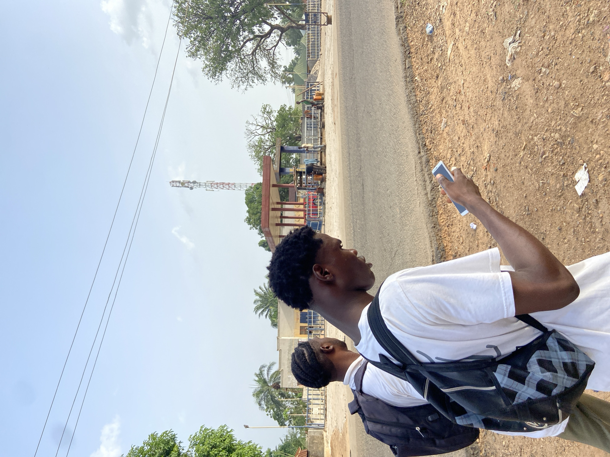 Two young men are depicted walking outdoors on a sunny day in Mararaba, Nigeria. The image is taken from a slightly elevated perspective, looking down and forward.
In the foreground, a young man with dark, short, curly hair is seen in profile, looking towards the left side of the frame. He wears a plain white t-shirt and a black backpack with a gray geometric pattern. His right hand, extended forward, holds a light-colored smartphone. Behind him, another young man is partially visible, also wearing a white t-shirt and a black backpack, with braided hair.
They are walking alongside a paved road, which occupies the middle left of the frame. To the right of the road is a wide, unpaved shoulder of dirt and gravel, with visible litter scattered across it.
The background features various elements consistent with a semi-urban or rural setting. Several trees are visible, including large deciduous trees and palm trees. Buildings with different roof colors, such as green and red-brown, can be seen behind the trees. A tall communication tower stands prominently in the distance on the left. Overhead, thin power lines stretch across the bright, light blue sky, which has scattered white clouds.
The scene suggests a casual outing or commute during daytime, under clear and bright weather conditions. No discernible text is visible on any objects or signs in the image.