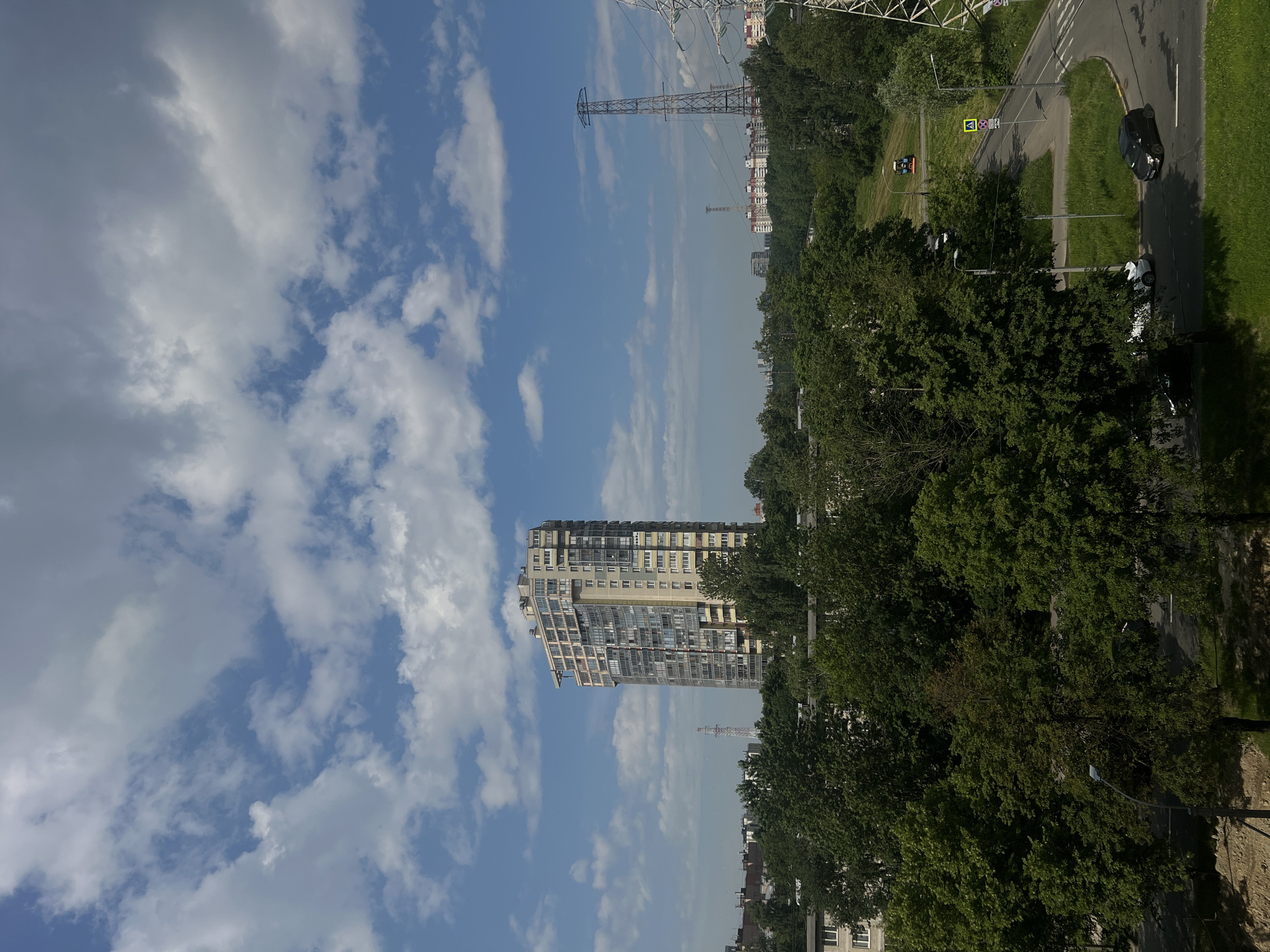 The image depicts a daytime scene in Petro-Slavyanka, Russia, characterized by a bright blue sky with scattered white clouds. In the mid-ground, a tall, modern apartment building with a light-colored facade and numerous windows stands prominently. It is surrounded by a dense canopy of green trees, suggesting a suburban or park-like setting within the city. In the distance, several other buildings and residential complexes are visible, along with high-voltage electricity pylons. A paved road curves through the foreground, with a few parked cars and a pedestrian crossing. A street lamp is also visible. The overall impression is a view from an elevated position overlooking a green, urban landscape under a clear sky.