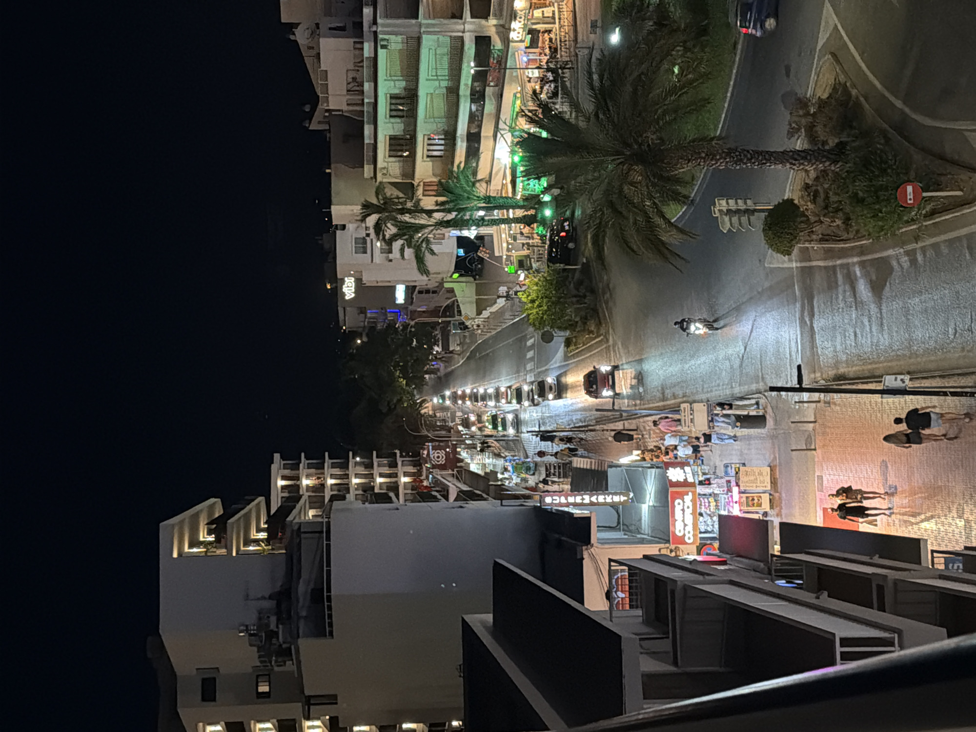 The image depicts a bustling street scene at night in Sant Antoni De Portmany, Spain. The scene is illuminated by the artificial lights of shops, restaurants, and streetlights, creating a vibrant and lively atmosphere.
The main subjects are the people walking along the street and sidewalks, suggesting a leisurely evening or a lively nightlife. There are also several vehicles, including cars and a motorcycle, indicating ongoing traffic. The architecture is characteristic of a Mediterranean coastal town, with multi-story buildings housing shops and establishments.
Notable details include palm trees, which are common in this region, adding to the tropical feel. The time of day is clearly night, as evidenced by the dark sky and artificial lighting. The presence of shops with open fronts and illuminated signs suggests that the area is a commercial and entertainment hub.
The visible text includes the word "vibi" on a sign, and another sign that reads "CENTRO COMERCIAL" which translates to "shopping center" in Spanish. These textual cues reinforce the location's nature as a place with shops and commercial activity. The overall emotion conveyed is one of energy and activity, typical of a popular tourist destination at night. The weather appears to be clear, as there are no indications of rain or adverse conditions.