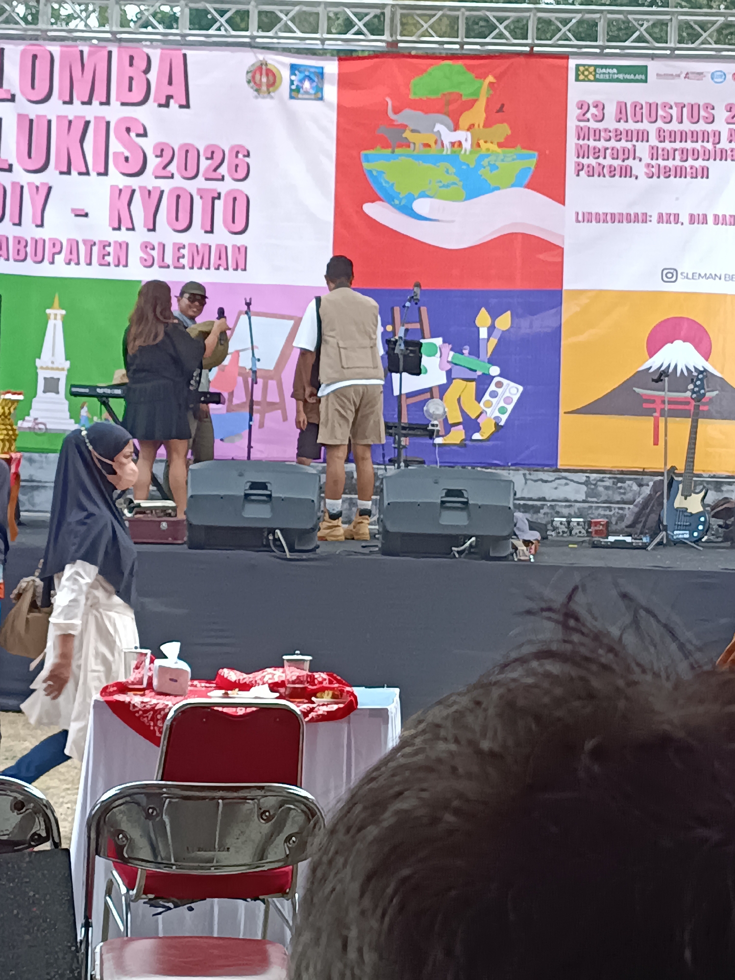 The image captures an outdoor event, likely a festival or competition, set against a backdrop of large banners. The banner on the left prominently displays text in pink letters that reads "LOMBA LUKIS 2026 DIY - KYOTO KABUPATEN SLEMAN," indicating a painting competition related to Kyoto, Japan, scheduled for 2026 in Sleman Regency. This suggests the event is located in Sleman Regency, Indonesia.
In the mid-ground, a stage area is visible. Several people are on or near the stage. To the left, two individuals are standing near a keyboard; one appears to be a woman in a black dress, and the other, possibly a man, is holding a microphone. Further to the right, a man in a tan vest and shorts stands near some musical equipment. A woman in a black hijab and white outfit is in the foreground, walking towards the right side of the frame. She is wearing a light-colored face mask.
Behind the stage, several large banners are displayed. One banner features an illustration of a hand holding a globe with various animals on it, above text that includes "23 AGUSTUS 201" suggesting a date. Another banner on the right depicts Mount Fuji with a red torii gate. Musical instruments, including a guitar, and speakers are also present on the stage area. In the lower foreground, a table covered with a red and white patterned cloth is set with cups and a tissue box, with red chairs placed in front of it. The overall environment appears to be an open-air venue, possibly a park or plaza, with a stage set up for performances or presentations.
Given the text on the banner, the event is located in Kabupaten Sleman, Indonesia. Hargobinangun is a village within the Cangkringan sub-district of Sleman Regency, Yogyakarta, Indonesia, and could potentially be the specific location of this event given the mention of "Kabupaten Sleman" on the banner.
