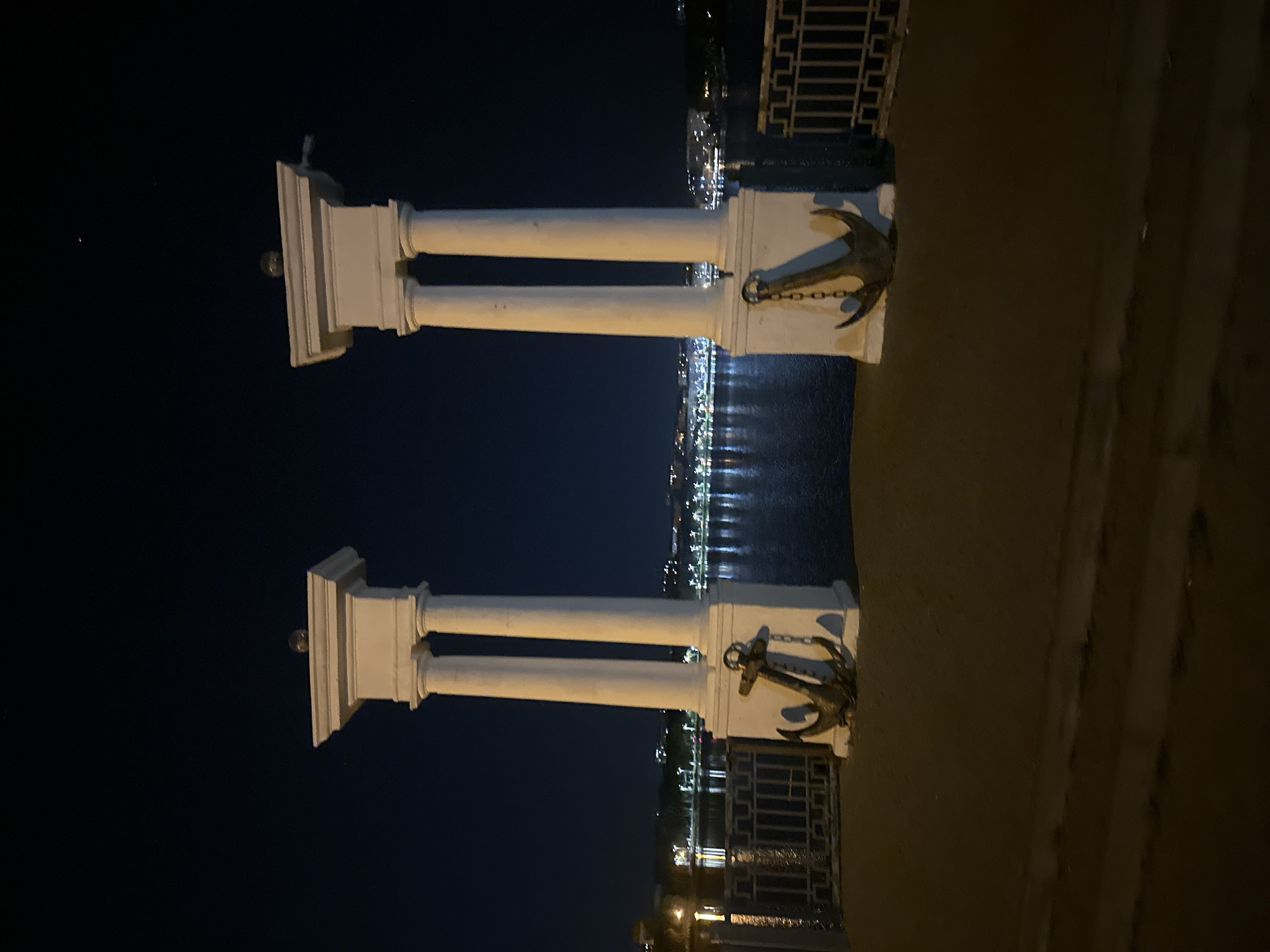 The image shows a nighttime view of a promenade along a body of water in Ternopil, Ukraine. Two decorative classical-style columns, topped with spherical lights, stand prominently on the left and right sides of the foreground. Attached to the base of each column is a large, antique-style anchor with a visible chain. A metal railing runs along the edge of the promenade, separating it from the water. Across the water, the lights of the city are visible, reflecting on the surface. The sky is dark blue, indicating nighttime. The ground in the foreground appears to be paved.