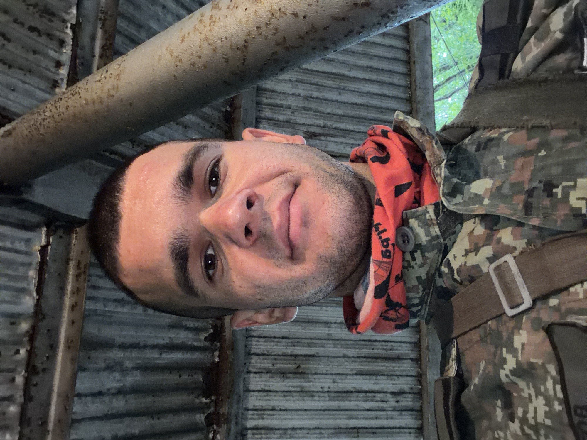 A young adult male with short dark hair and a shaved head is pictured in a selfie, looking directly at the camera. He is wearing a camouflage jacket and an orange neck gaiter with a black pattern. The background consists of a corrugated metal roof and a large, weathered cylindrical pillar. The overall environment appears to be an outdoor shelter or structure, possibly in a rural or natural setting, given the glimpse of green foliage visible in the upper right portion of the image. The scene is lit by natural light, casting subtle shadows. The man's facial expression is neutral.