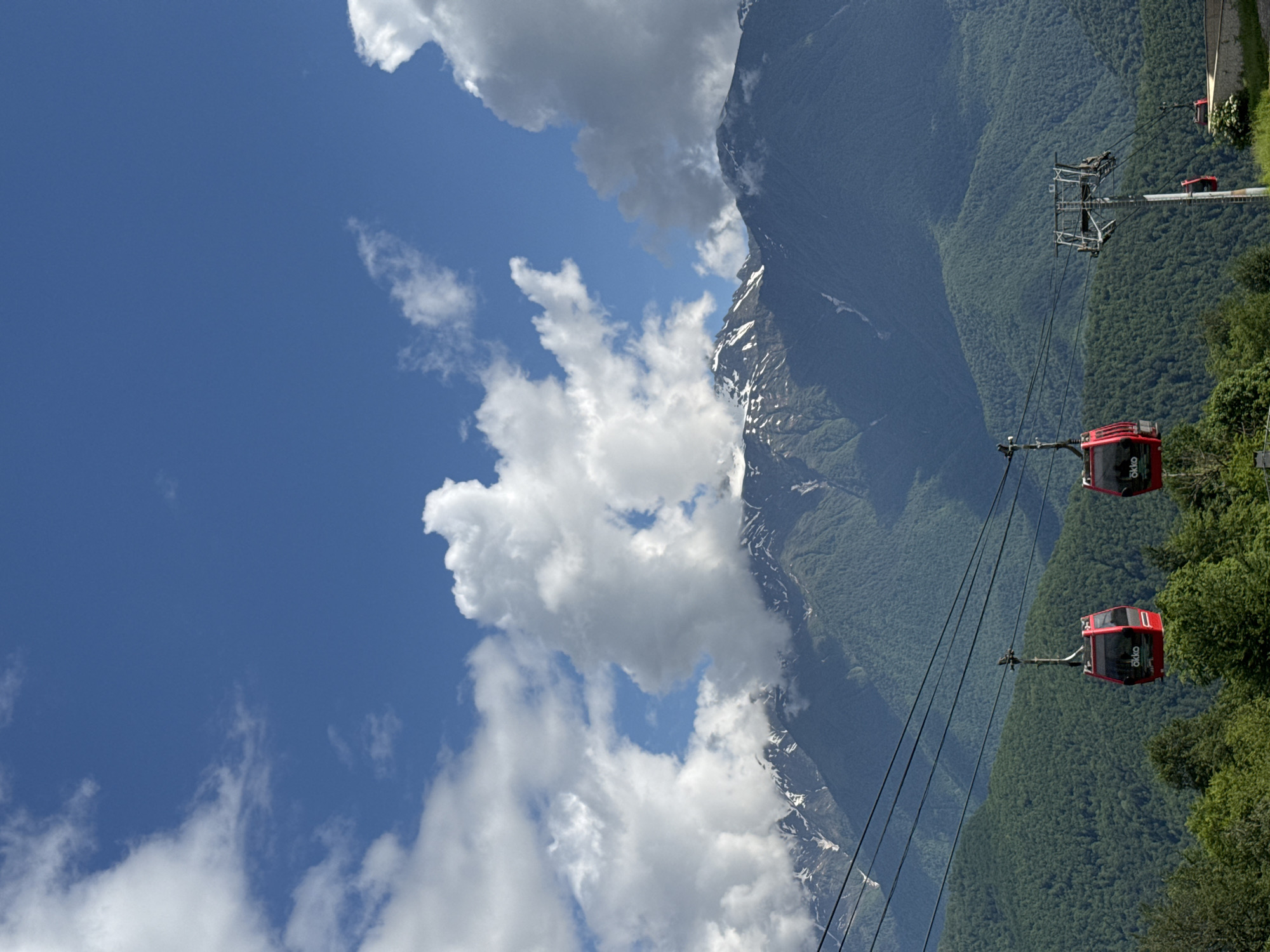 This is a full-frame, vertical shot of a mountainous landscape on a clear, sunny day with scattered white clouds in a bright blue sky. In the foreground, two red cable cars with the "Okko" logo are visible, suspended from black cables that stretch across the frame. The cable cars appear to be in motion. The middle ground is dominated by lush green, densely forested mountains. In the background, towering snow-capped mountains rise above the tree line, with patches of snow visible on their peaks and slopes. The sky takes up the top third of the image, with the mountains and cable cars occupying the lower two-thirds. The scene depicts a cable car system operating in a mountainous region near Estosadok, Russia, likely during warmer months given the green foliage. There are no people visible in the image.
