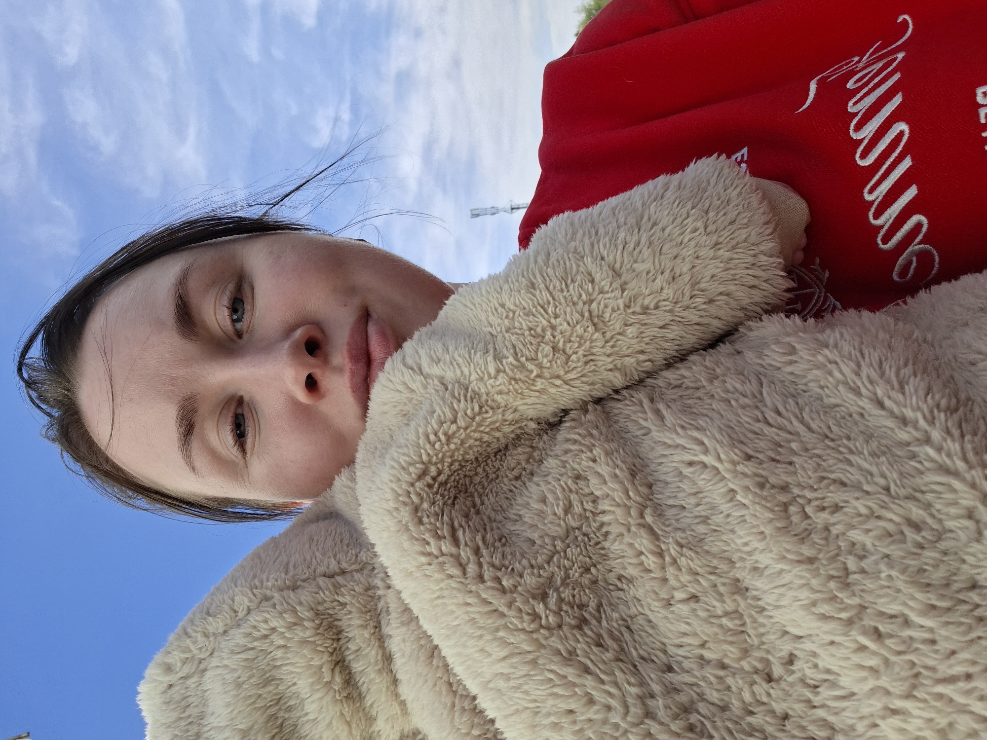 A close-up, tilted shot shows a woman with fair skin and dark hair. Her face, partially framed by the clear blue sky with scattered clouds, is tilted slightly down towards the camera. The woman is wearing a fluffy, light-beige jacket, the texture of which is prominently displayed. Parts of a red garment are visible behind the jacket. The image was likely taken in an urban environment, as suggested by the presence of a communication tower or similar structure in the far background. The precise location and city are not identifiable.