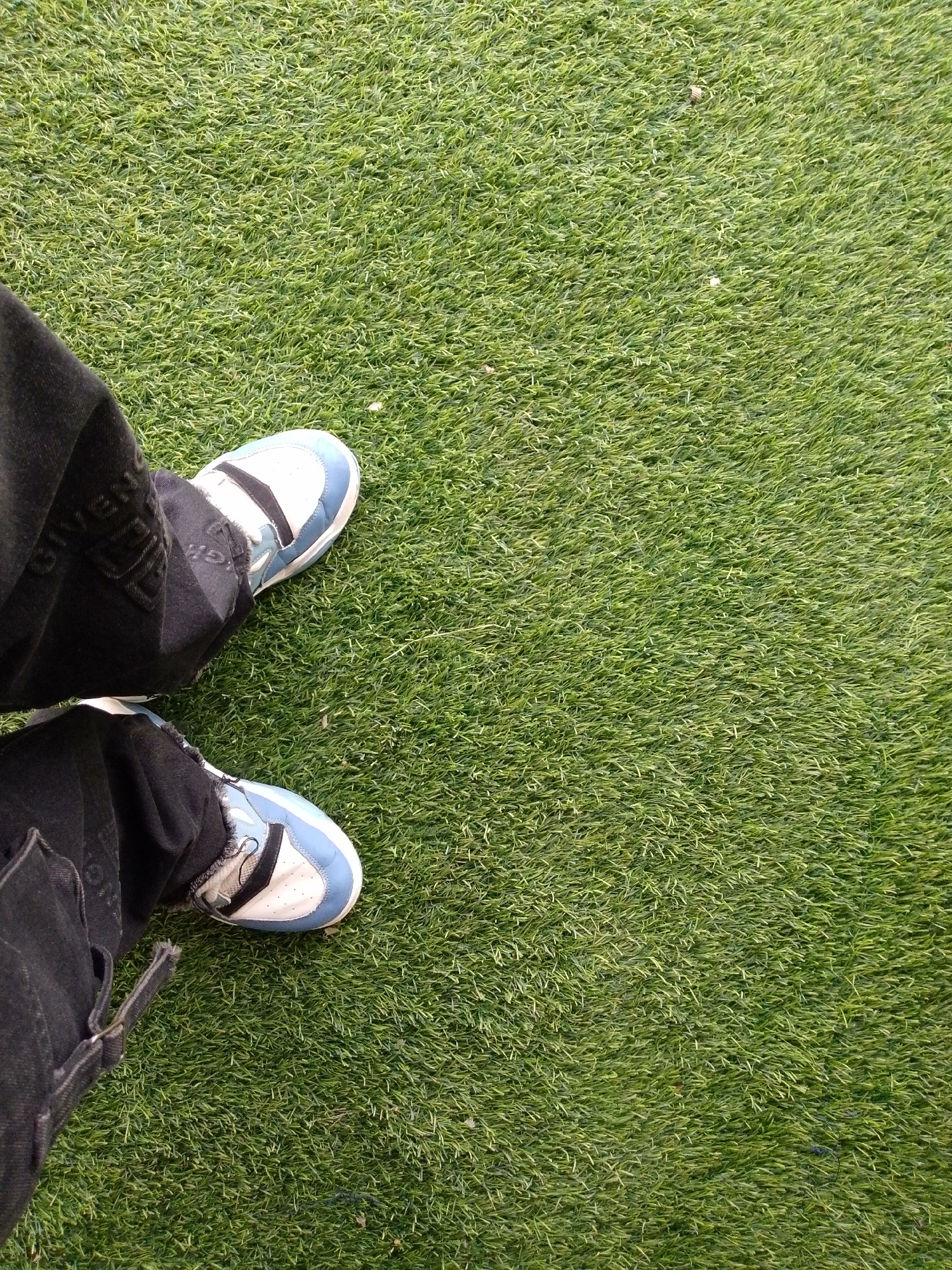 A first-person perspective shot shows a pair of black pants and white and blue sneakers standing on artificial green turf. The pants are slightly frayed at the bottom. The sneakers are laced and have visible logos. The artificial turf appears to be a consistent, vibrant green with no visible debris or damage. The lighting suggests an outdoor environment, possibly during the day. The location is identified as Barikin Jirga Summa, Nigeria.