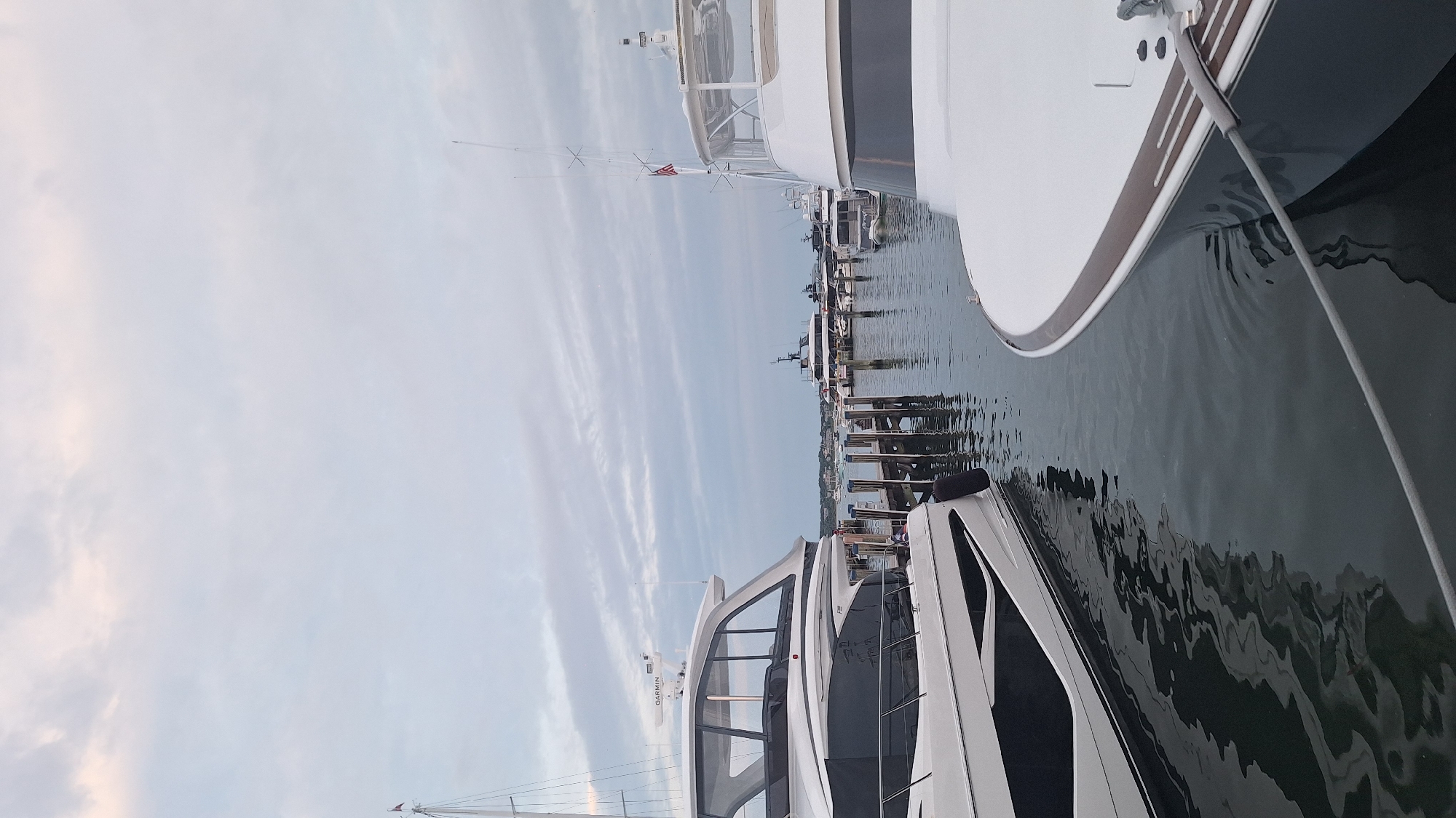 An overcast, early morning scene depicts a harbor in Nantucket, Massachusetts, United States of America. The foreground is dominated by the bow of a large, white luxury yacht, tethered by a thick grey rope to a dock that is not fully visible. To the left, the stern of another sleek, white yacht with black accents is moored, its reflection creating an abstract pattern on the dark, rippling water. In the middle ground, a wooden dock with several pilings extends into the harbor, with smaller boats and larger vessels moored beyond. In the background, a distant shoreline with buildings and trees is visible under a cloudy sky. The overall impression is one of quiet luxury and maritime activity.
