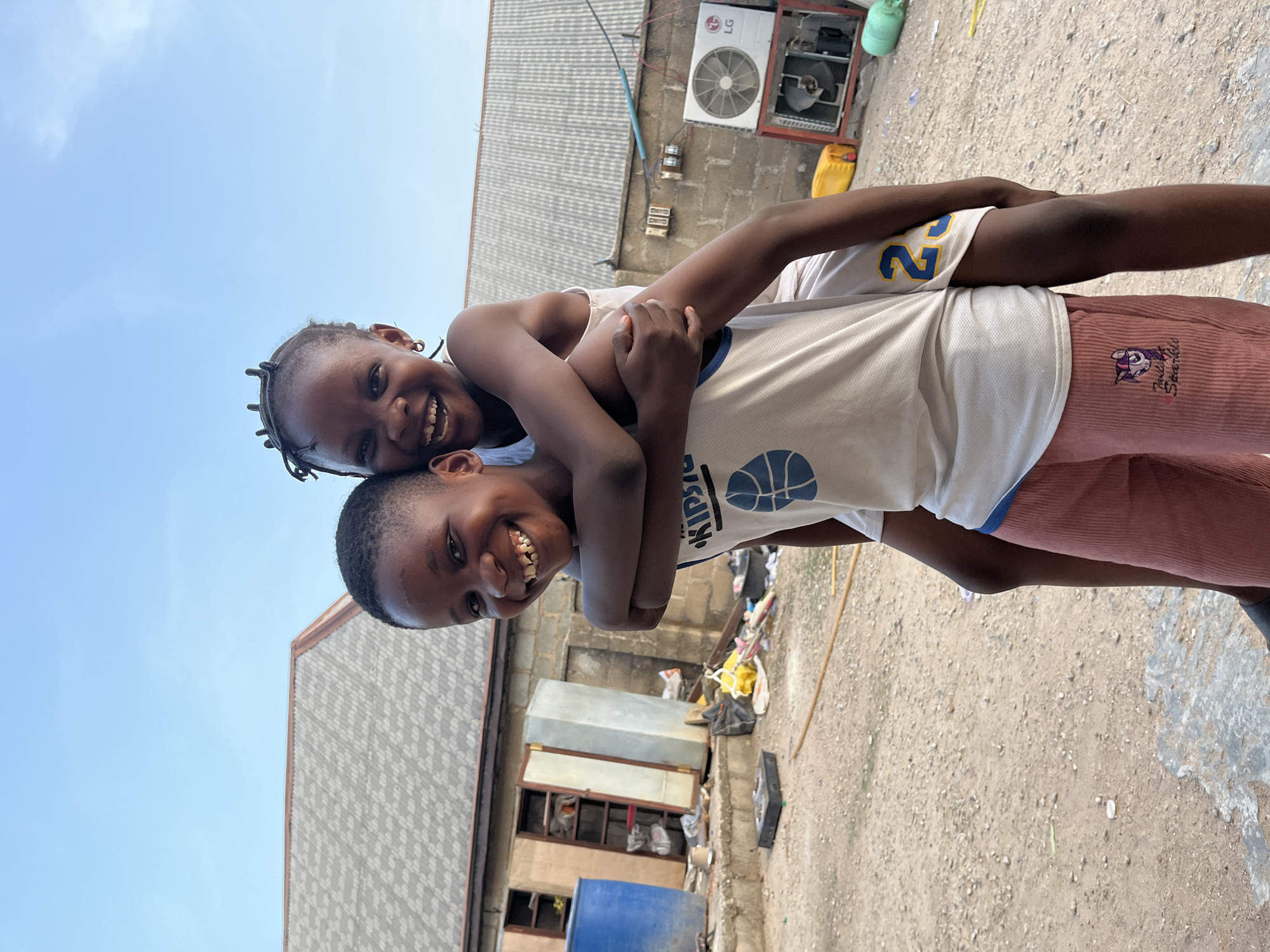 Two children are photographed outside in Iddah, Nigeria. One child carries the other on their back. Both children are smiling broadly. The child on the bottom is wearing a light-colored basketball jersey and maroon pants with a cartoon character on the leg. The child on top is wearing a light-colored tank top. The background shows corrugated metal rooftops, a brick building with an air conditioning unit, and a dirt ground with scattered trash and debris. The sky is visible in the upper left corner.