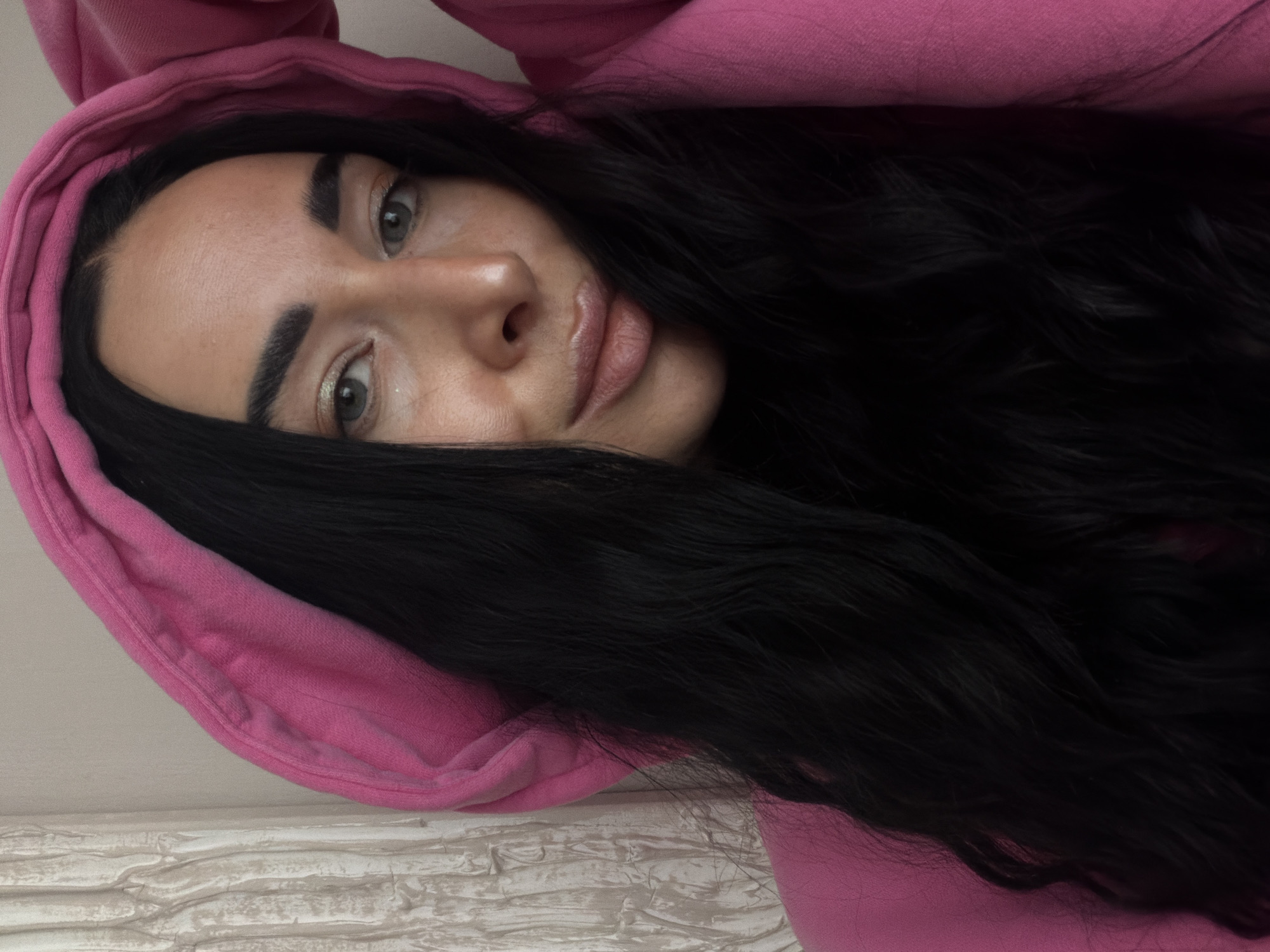A woman with long, dark hair is wearing a pink hoodie. She is looking at the camera. The background is a white wall with a textured surface.